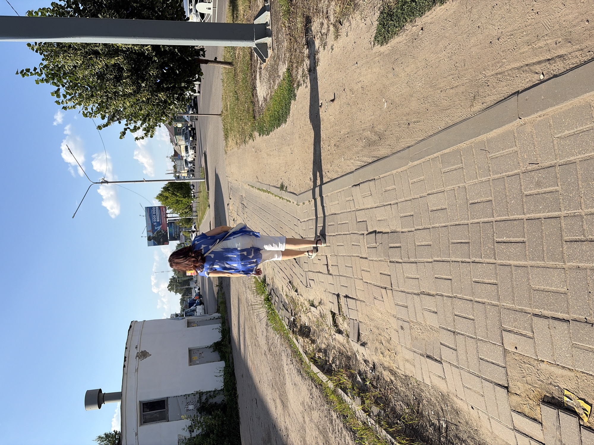 The image depicts a sunny day on a paved pedestrian walkway in Tambov, Russia. A woman with dark hair, wearing a blue and white patterned blouse and white capri pants, walks away from the camera towards the right side of the frame. She carries a white strap bag over her left shoulder. The walkway is made of grey interlocking pavers, some of which appear uneven or damaged. Adjacent to the walkway is a sandy area with patches of dry grass. In the background, a white cylindrical building with two doors and a window is visible on the left, with graffiti on its walls. Further back, a street with several cars driving on it is seen. A large billboard advertising a lottery is also present. On the right side of the frame, a tall grey pole, likely a lamppost, stands next to a leafy green tree. The sky is clear blue with a few white clouds. The overall scene suggests an urban or suburban environment.
