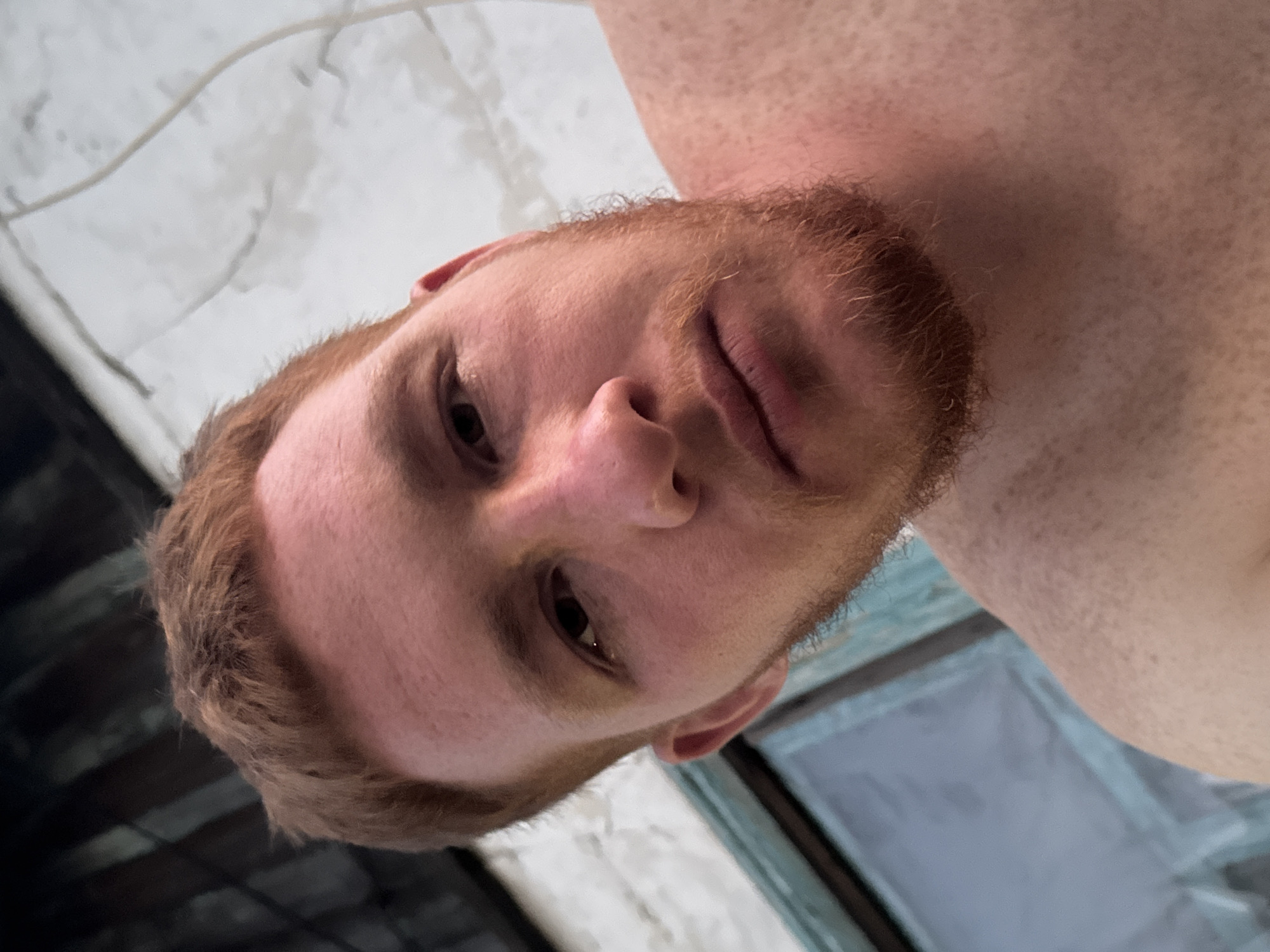 A young adult male with reddish-brown hair and a beard is pictured in a close-up, selfie-style shot. He is shirtless, and his skin has a fair complexion with freckles. The background is out of focus, showing what appears to be a wall with some light-colored texture, possibly painted concrete or stucco, and a faint wire or pipe running vertically. To the left of the frame, a portion of a window with a light blue frame is visible. The lighting suggests it is daytime, possibly overcast or late afternoon, casting soft shadows. The subject's expression is neutral, and his gaze is directed towards the camera. The overall environment appears to be outdoors, with the building providing a backdrop.
