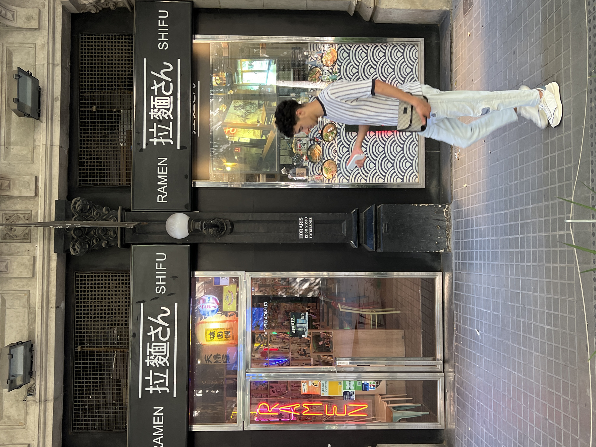 A young man walks past a ramen restaurant in Barcelona, Spain. The restaurant, named "Ramen Shifu", has a black facade with a large sign displaying Japanese characters and the word "RAMEN" in English. The entrance is made of glass, with neon signs inside. The man is wearing a striped t-shirt, ripped jeans, and white sneakers, and is carrying a small bag over his shoulder. The street in front of the restaurant is paved with gray tiles. The man is captured mid-stride, looking to his right as he walks. The lighting suggests it is daytime, possibly late afternoon, with shadows cast on the ground. The overall scene depicts a casual urban street view.