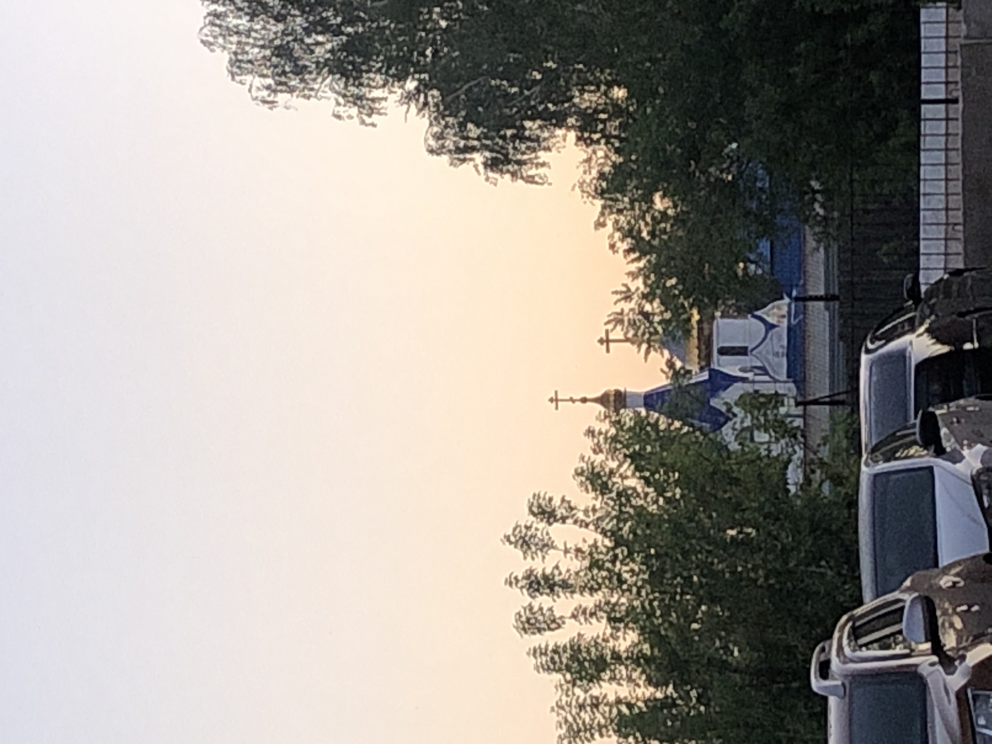 The image displays a serene outdoor scene, likely at dawn or dusk, indicated by the soft, warm gradient of the sky from pale white to a light orange/yellow. The dominant subject is a religious building, resembling an Eastern Orthodox church or chapel, positioned slightly to the left of the center. It features characteristic blue and white domes topped with golden crosses.
Lush green trees partially frame and obscure the view of the church, with foliage visible in the foreground, midground, and background. On the right side of the frame, parts of several parked vehicles, possibly cars or trucks, are visible, suggesting an urban or suburban setting. A light-colored building or wall structure is also seen on the far right. The overall impression is one of calm, with no human activity or specific events taking place. The weather appears clear with no clouds in the sky. No text is visible in the image.