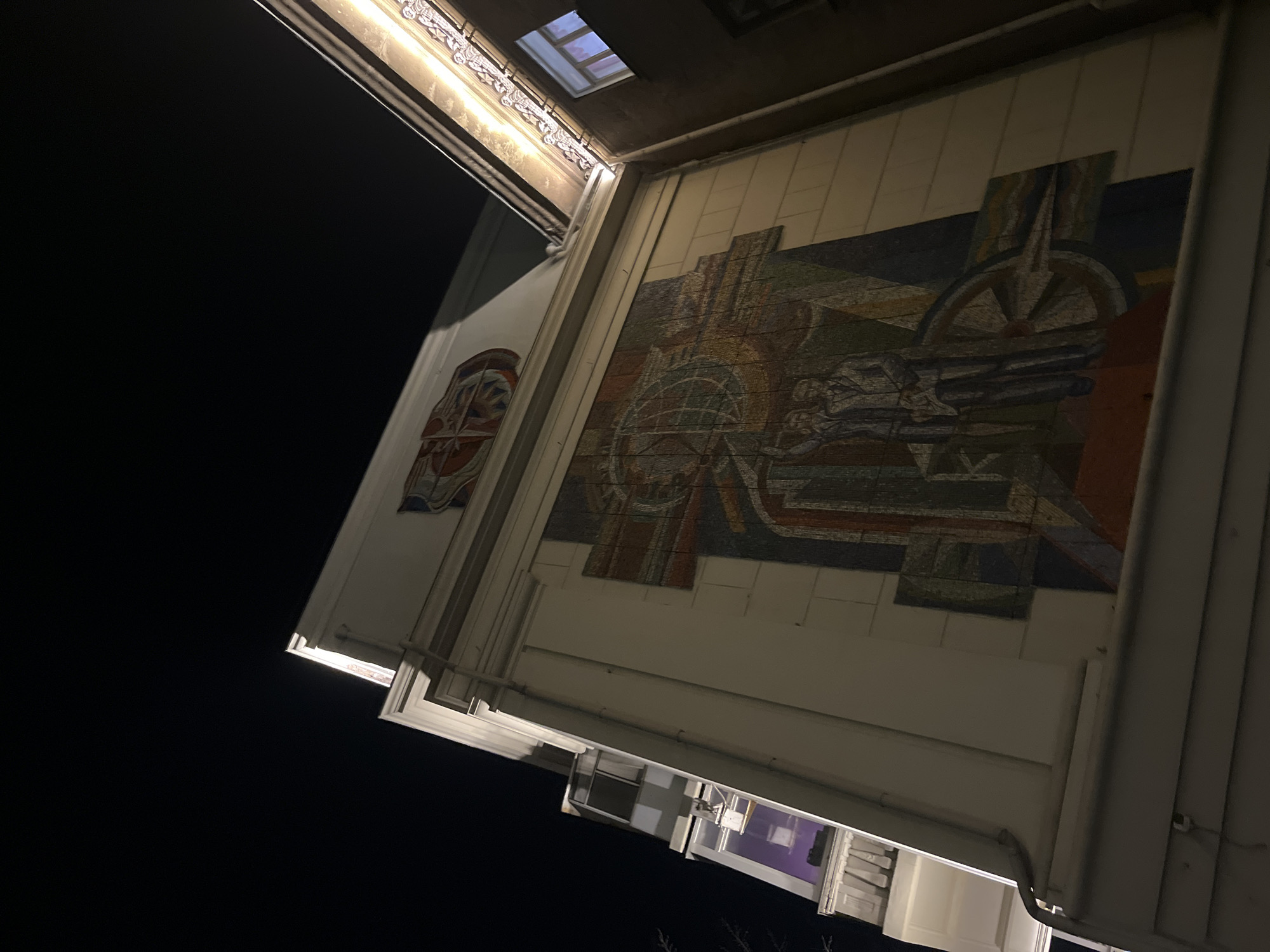 The image shows a building with a mosaic mural, likely a Soviet-era design, located in Krasnoyarsk, Russia. The mural features abstract shapes and possibly a globe or similar object. The building has a light-colored facade and is viewed at an extreme angle from below, with a dark sky visible on the left side. Other architectural details like windows and decorative moldings are visible on the building. There are no people visible in the image. The time appears to be night.