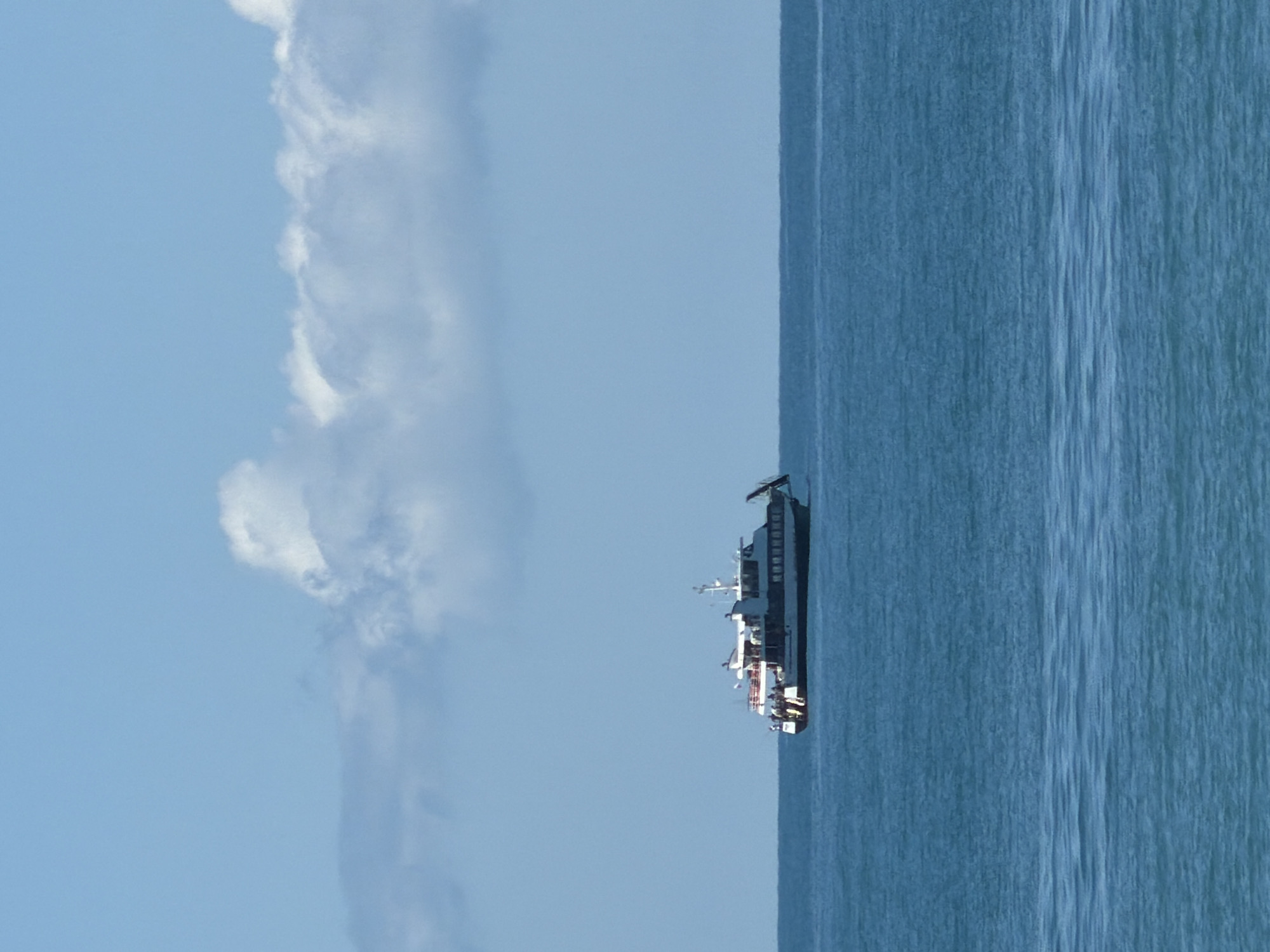 A medium shot captures a ferry boat sailing on the open sea under a bright blue sky with scattered white clouds. The ferry, predominantly white with dark trim, has multiple decks and appears to be carrying passengers, with several figures visible on the upper deck. The boat is positioned slightly to the left of the frame, cutting across the horizontal expanse of the water. The sea is a deep blue, showing slight ripples on its surface. The horizon line is clearly defined where the sea meets the sky. The image does not depict any elements specific to Sochi, Russia; it is a generic seascape.