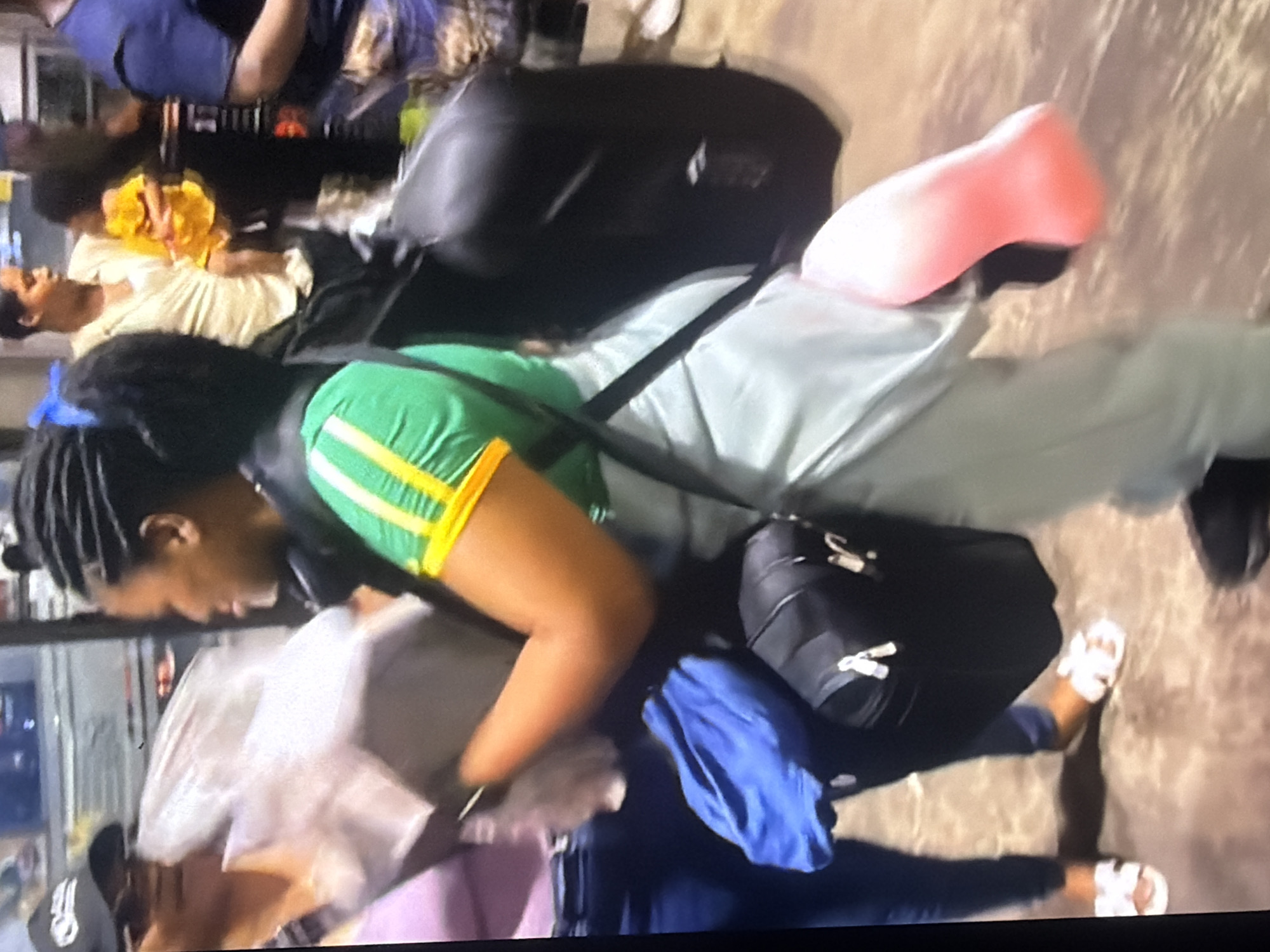 In Bukuru, Nigeria, a person stands in a well-lit indoor area, possibly an airport terminal or a transport hub, given the presence of luggage. The individual is facing right, with their left side partially obscured by a large, black duffel bag. They are wearing a green shirt with yellow and white striped trim, and light blue pants. A pink prosthetic leg is visible, attached to their right leg and extending downwards. The person is carrying several items, including a blue cloth bag slung over their left shoulder and a large, opaque plastic bag held in their hands, appearing to contain various objects. To the left, another person, wearing a black baseball cap and a lavender shirt, is partially visible, holding a similar opaque plastic bag. The ground is a textured, mottled brown surface. In the background, other people and luggage are discernible, suggesting a public space with ongoing movement.