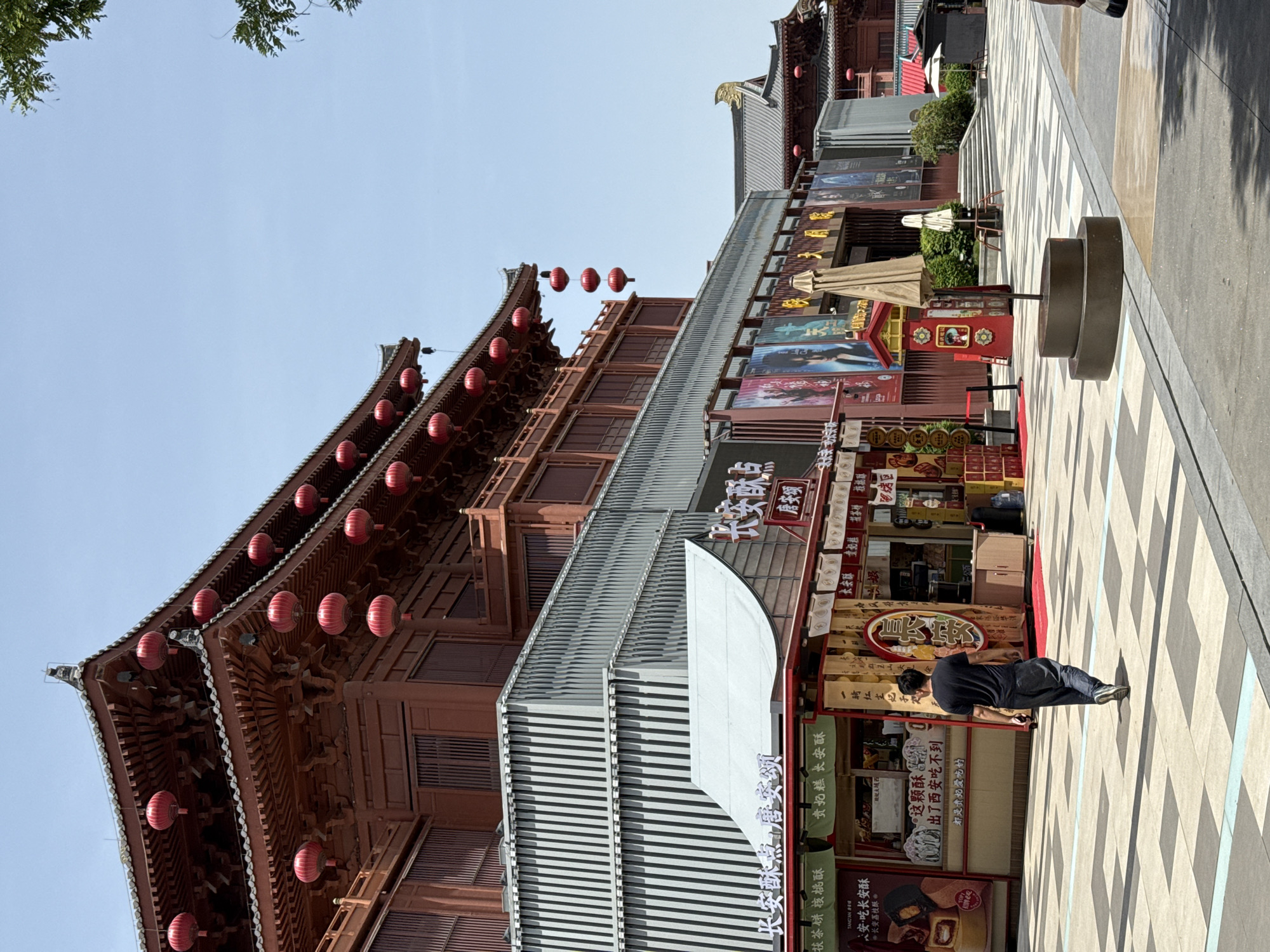 The image depicts a street scene in Yanta District, China, featuring traditional Chinese architecture adorned with red lanterns. A person dressed in black bends over, appearing to examine or interact with a storefront. The storefronts display advertisements and signage in Chinese script, specifically for "Chang'an Su Dian Tang An Song," indicating a local specialty shop, possibly selling pastries and teas like "Fucha Cake Walnut Cake." The storefronts also include posters and decorations that suggest a commercial area catering to tourists. The ground is paved with a checkered pattern, and there are street fixtures, including a cylindrical structure, visible in the foreground. The architecture and signage indicate a cultural or historical district within the city.