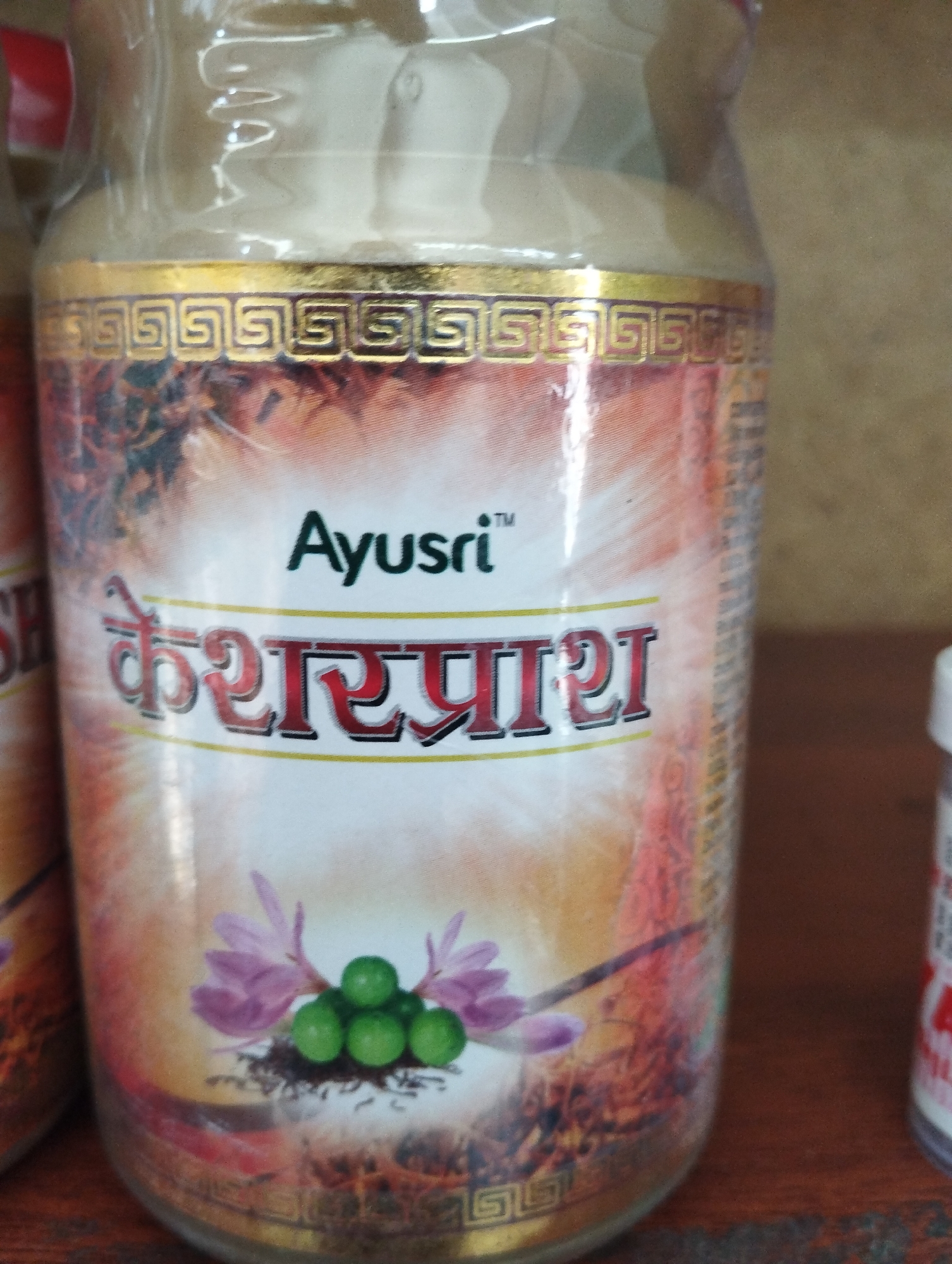 The image shows a close-up of a glass jar containing a powder. The jar has a golden decorative band with a Greek key pattern near the top. Below the band, there is text that reads "Ayusri" in English and then "केशरप्राश" in Hindi. There are also images of purple flowers and green berries on the jar. The jar appears to be sitting on a wooden surface. The background is blurred, but it appears to be a wall or shelf. The lighting is soft and diffused. The overall impression is that of a product for sale, likely in a market or store.