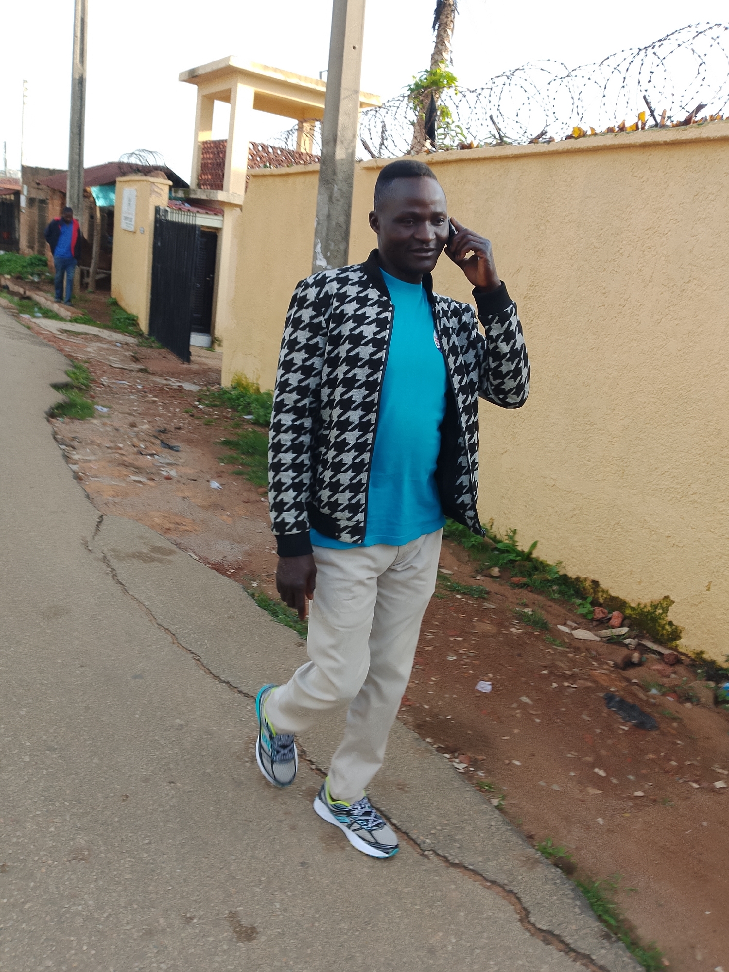 In Jos, Nigeria, a Black man in his late 20s to early 30s is walking down a cracked concrete sidewalk next to a dirt path. He is wearing a black and white houndstooth patterned jacket over a bright blue t-shirt and light-colored pants. His right hand is holding a cell phone to his ear. He is wearing grey and blue sneakers. Behind him, a yellow building with a darker yellow second story is visible, bordered by barbed wire. Further in the background, another building with a red roof and a person in a blue jacket and jeans can be seen. The environment appears to be an urban street with some overgrown vegetation and debris.
