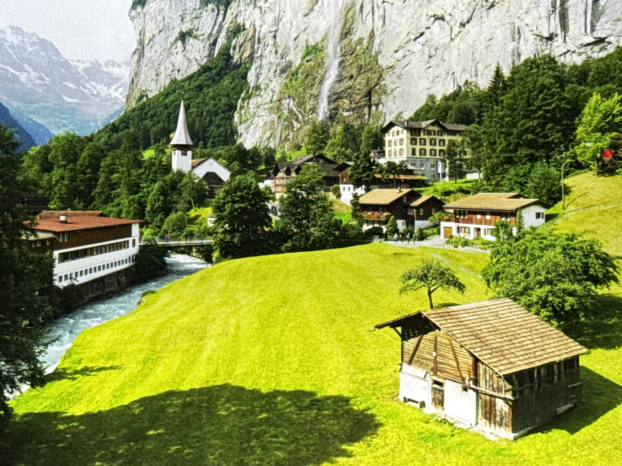This is an image of a scenic village nestled in a valley with a dramatic waterfall cascading down a sheer cliff face. The village consists of several traditional chalets and a church with a steeple, surrounded by lush green trees and rolling hills. A river flows through the valley, and a bridge crosses it. The image captures a bright, sunny day, with shadows indicating strong sunlight. This scene does not depict the city of Burgas, Bulgaria, but rather a mountainous, European alpine landscape.