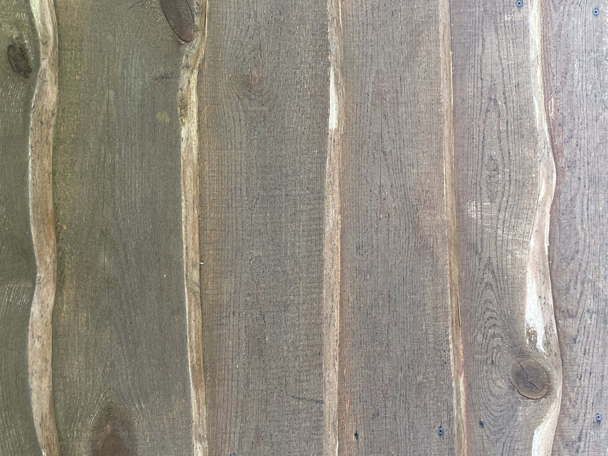 The image shows a close-up, vertical view of weathered wooden planks, likely part of a building or fence. The planks are horizontally oriented and vary slightly in color and texture, ranging from dark gray to brown. They are joined together, creating a textured surface with visible wood grain, knots, and occasional nail heads. The overall appearance suggests outdoor exposure to the elements. The specific location in Norviliškės, Lithuania, is not discernible from the image itself.