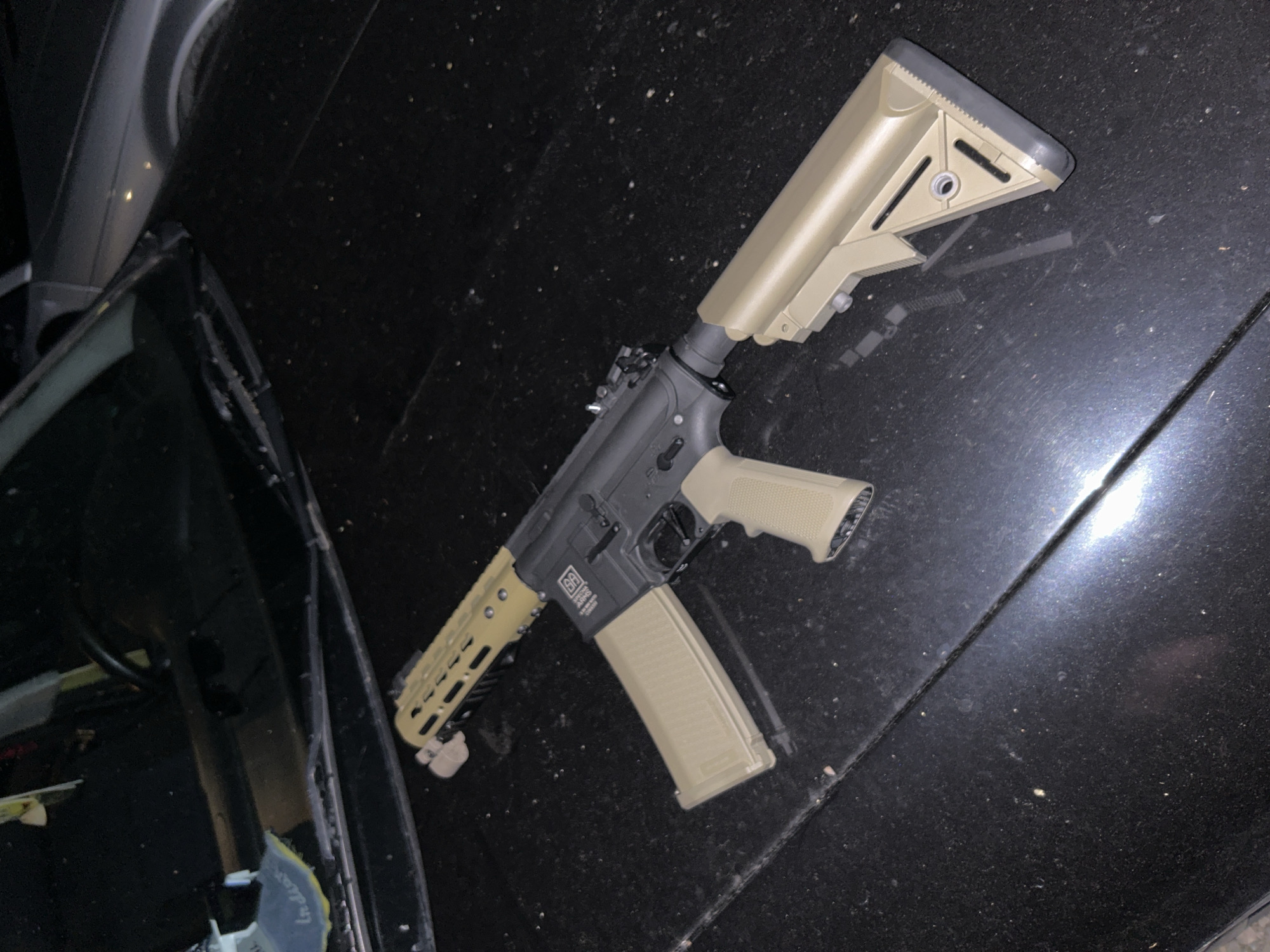 The image depicts a tan and black AR-15 style rifle resting on the hood of a black vehicle. The rifle is oriented diagonally across the frame. The surrounding environment is indistinct except for the car hood and part of a windshield. The location is not identifiable from the image.