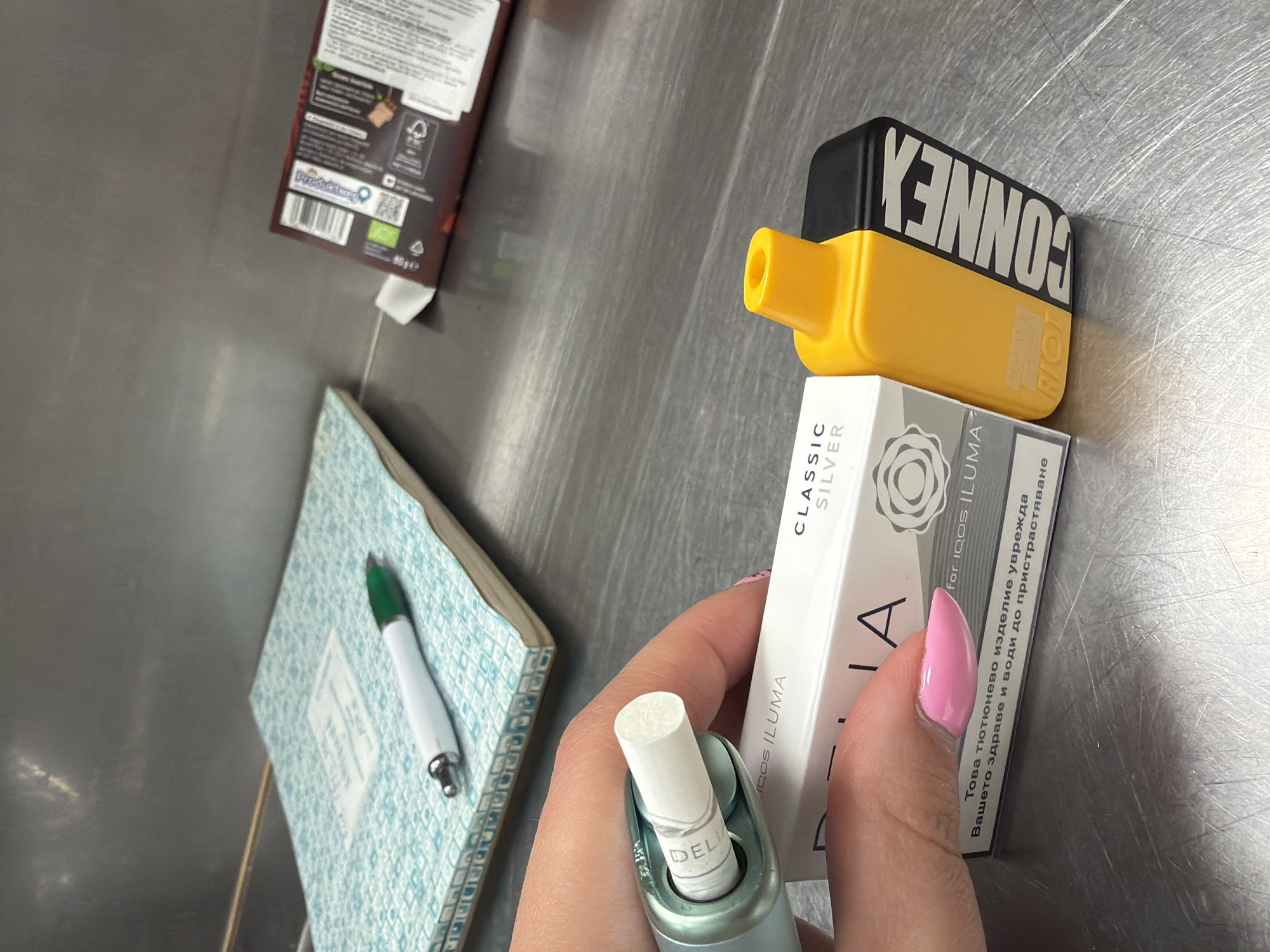 The image presents an overhead view of various items arranged on a brushed metal surface, likely a table or counter, in an indoor setting. In the central foreground, a person's hand, featuring a pink polished thumbnail, holds a light green and silver heated tobacco device. A white tobacco stick, partially visible with the brand "DELIA," is inserted into the device.
Adjacent to the hand and device is a white rectangular box of tobacco sticks, labeled "CLASSIC SILVER for IQOS ILUMA." Prominently displayed on this box is a health warning in Bulgarian Cyrillic: "Това тютюнево изделие уврежда Вашето здраве и води до пристрастяване," which translates to "This tobacco product harms your health and leads to addiction," indicating the location is Plovdiv, Bulgaria.
Stacked on top of this white box is a yellow and black vaping device, which has the brand "CONNEX RIO" printed on it in inverted text. In the background, to the left, an open notebook with a light blue and white patterned cover rests on the surface, with a green and white pen placed on its pages. Further in the upper left background, a brown cardboard box, possibly containing a food item, is visible with "Produitwao" and "80ge" (80 grams) printed on it, along with a QR code and an organic certification logo. The arrangement suggests a casual break or a collection of personal effects.