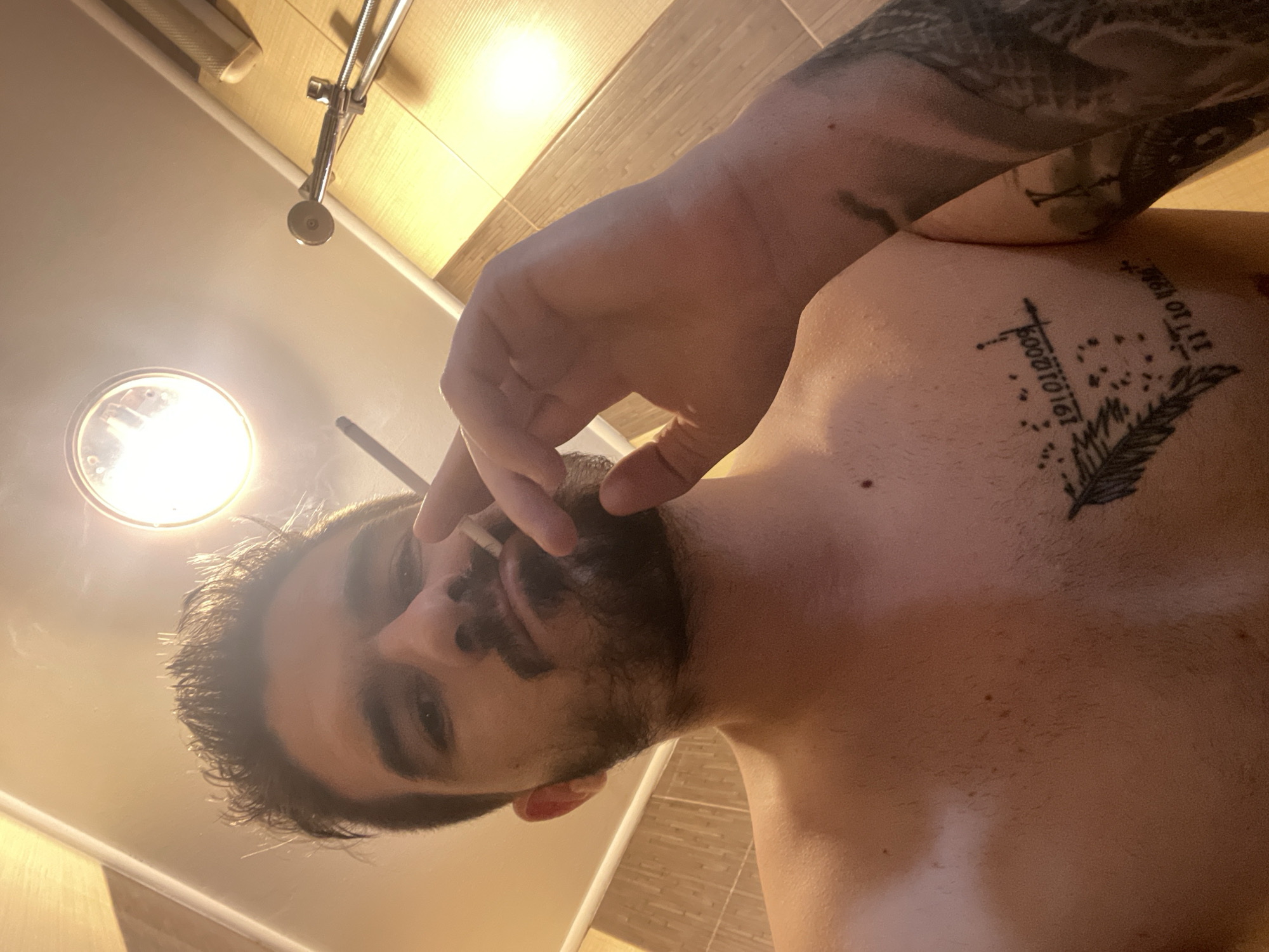 This is a low-angle, tilted photograph of a man in what appears to be a bathroom. The man is shirtless, with dark hair and a beard or stubble. He is holding a lit cigarette between his fingers, with smoke visible around his face. His gaze is directed towards the camera.
**Subjects:** The primary subject is a man. Other visible objects include a cigarette, a bright circular light fixture on the ceiling, and a metallic shower head or fixture attached to a tiled wall.
**Activity or event taking place:** The man is smoking a cigarette while being photographed, likely a selfie given the angle.
**Notable details:** The image is taken from a very low perspective, looking upwards, and is significantly tilted. The setting is an indoor space, strongly suggested to be a bathroom by the presence of the shower fixture and light-colored tiled walls. A prominent, bright circular light fixture illuminates the scene from above. The man has extensive tattoos: a dark, intricate sleeve tattoo on his right arm, and chest tattoos featuring a feather design, abstract symbols, and what appear to be dates.
**Visible text:** On his chest, among the tattoos, the dates "19.01.2005" and "11.10.1988" are clearly visible, along with other unidentifiable symbols.