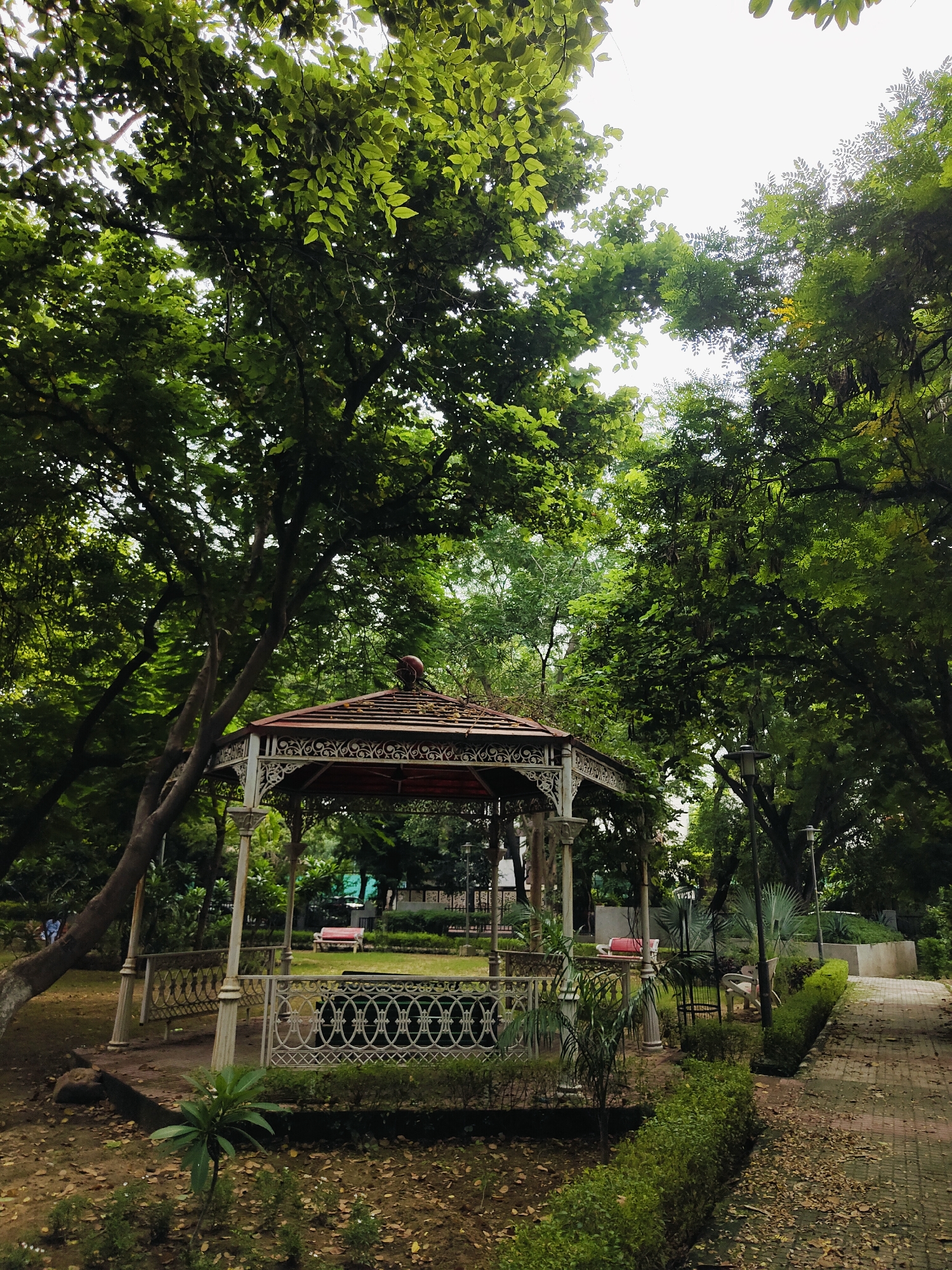 The image depicts a park with a white gazebo in the center, surrounded by lush green trees and foliage. The gazebo has a red roof and intricate white railings. A paved pathway lined with manicured bushes leads to the right of the gazebo. In the background, a red bench is visible on a grassy area. The scene appears to be a serene public space, likely in a park in Gurgaon, India. The overall ambiance is peaceful and natural, with the canopy of trees filtering the daylight.