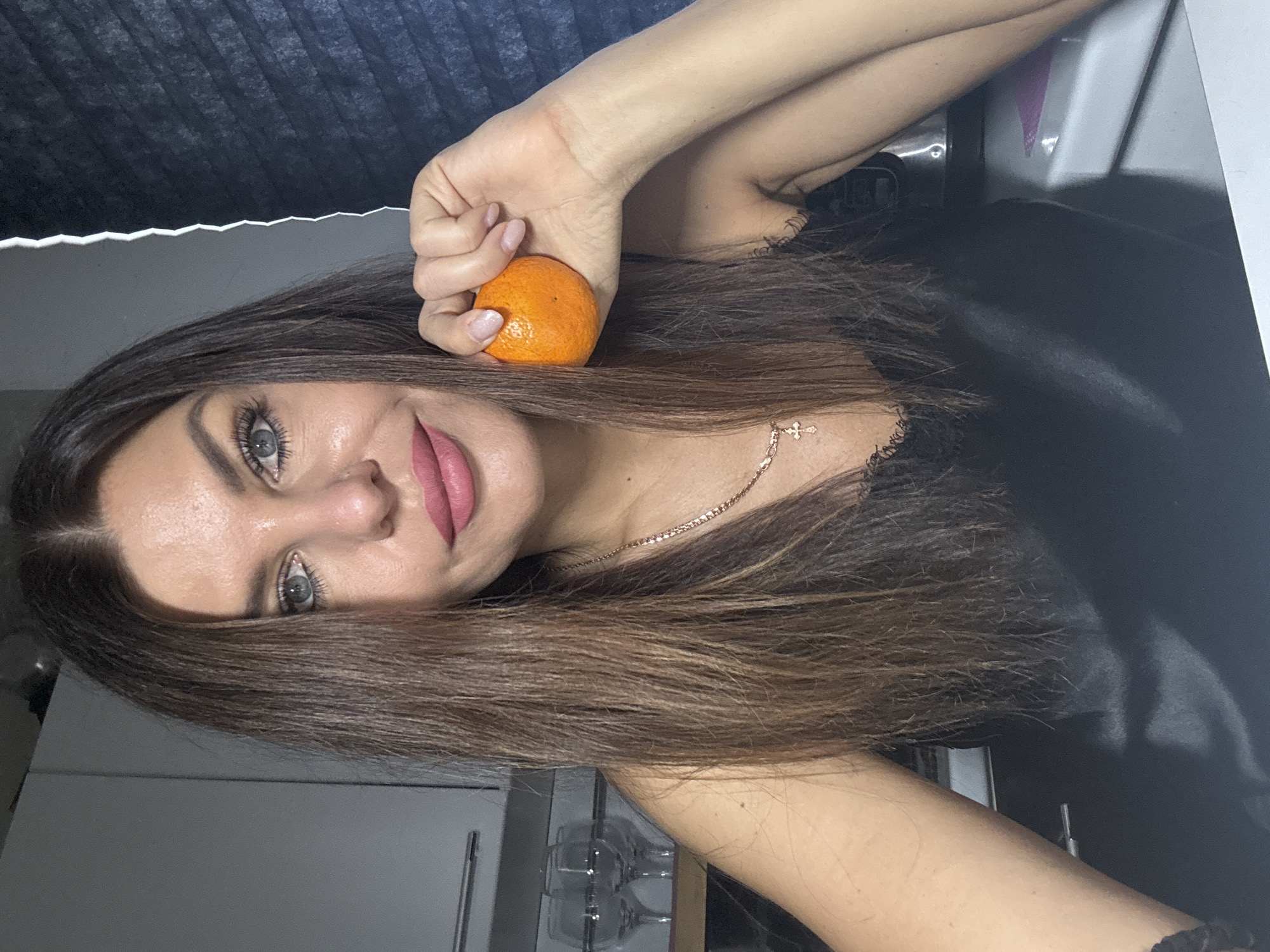 A young woman with long brown hair, light gray eyes, and wearing a thin gold necklace is holding a small orange citrus fruit near her forehead. She is wearing a black top. The background appears to be an indoor setting with glimpses of kitchen cabinets and a window covering. There is no information in the image to determine whether the location is Chaikovsky, Russia.
