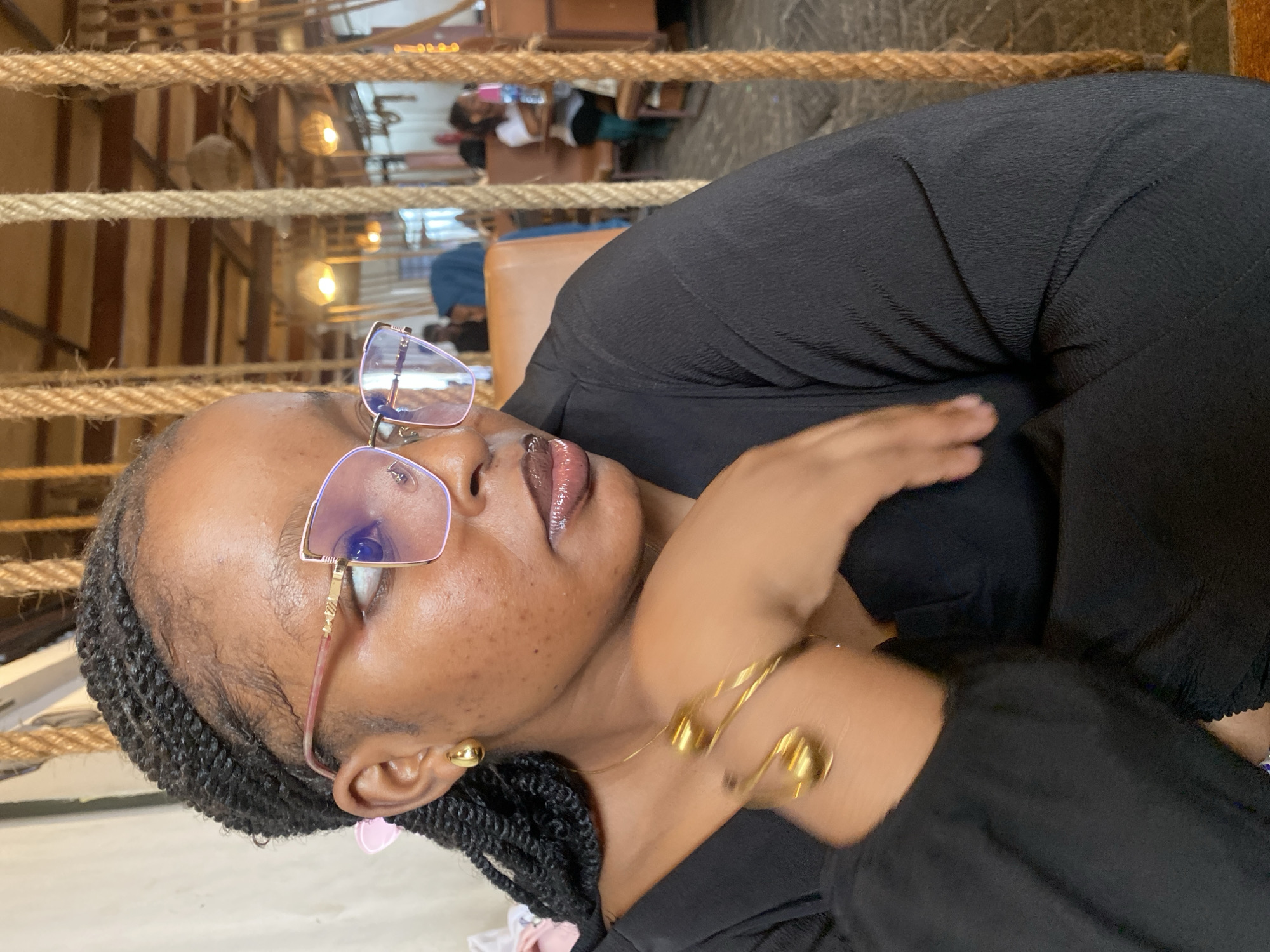 A person with brown skin is wearing eyeglasses and a black shirt. They have braided hair, a gold necklace, and gold earrings. The background features brown rope, wood paneling, and out-of-focus people. The location is in Dar es-Salaam, Tanzania.
