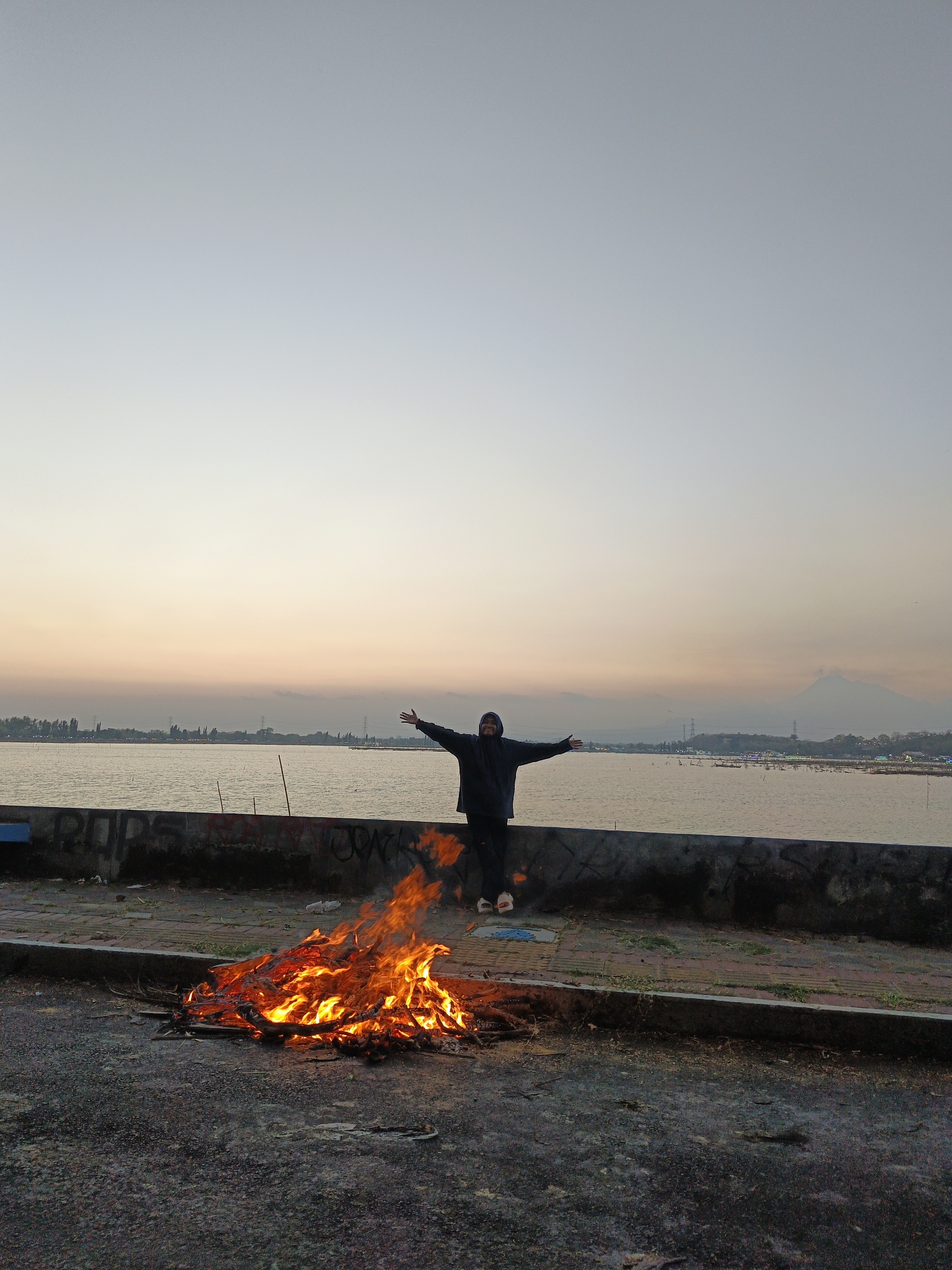 A lone individual stands with arms outstretched, silhouetted against a dusky sky, by a waterfront in Krakitan, Indonesia. In the foreground, a bonfire blazes on the asphalt. The person, wearing a dark hooded jacket and pants, is positioned on a paved walkway behind a low concrete barrier that borders a calm body of water. Beyond the water, a distant shoreline is visible with trees and structures. The sky exhibits a gradient of pale blue to soft orange hues, suggesting either sunrise or sunset. The overall scene captures a moment of solitary contemplation or celebration by the water's edge, with the fire providing a focal point of warmth and light.
