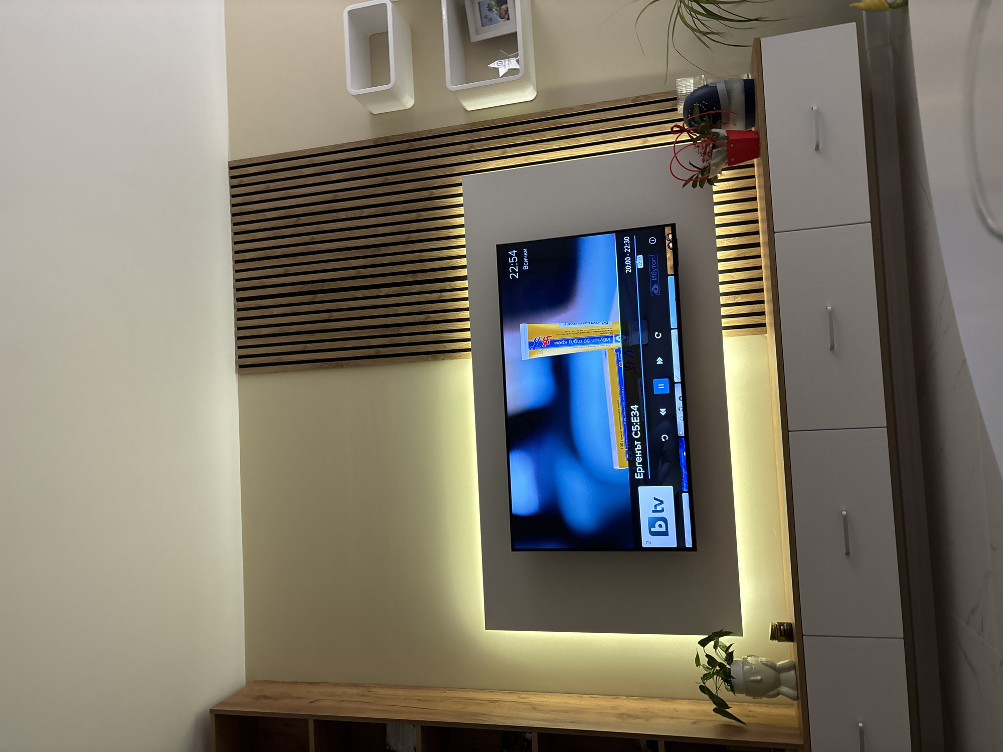 An interior shot captures a modern living room's entertainment area, tilted slightly to the right. A large flat-screen television is centrally mounted on a white rectangular panel, which is softly backlit by warm yellow LED lighting, creating a glowing frame. Behind this panel, and extending to the left, is a decorative slatted wooden panel, also illuminated from its edges.
The television screen displays a program from the Bulgarian channel "btv," identified by its logo. Visible text on the screen includes "Ергенът С5:E34," indicating "The Bachelor Season 5: Episode 34." An advertisement for "Ибутоп 50 mg/g крем" (Ibutop 50 mg/g cream) is partially visible. A digital clock reads "22:54," and a program guide shows a time slot from "20:00-22:30," with "22:13" highlighted, suggesting the current time, and the word "Ибутоп."
To the right of the TV, a tall, narrow white cabinet with two doors and silver handles stands. Above the wooden slatted panel, two white, open rectangular wall shelves are mounted, one containing a small framed picture. Below the television, a wooden media console with storage compartments supports a small white decorative planter holding a green plant. Another small plant in a white pot is visible on the console. Decorative items, including a red pot with artificial flowers and a blue vase, are placed on top of the right cabinet.
The walls are painted in a light cream color, complementing the light wood tones and white furniture, contributing to a warm and contemporary aesthetic. The prominent backlighting around the TV and slatted panel indicates an evening or nighttime setting.