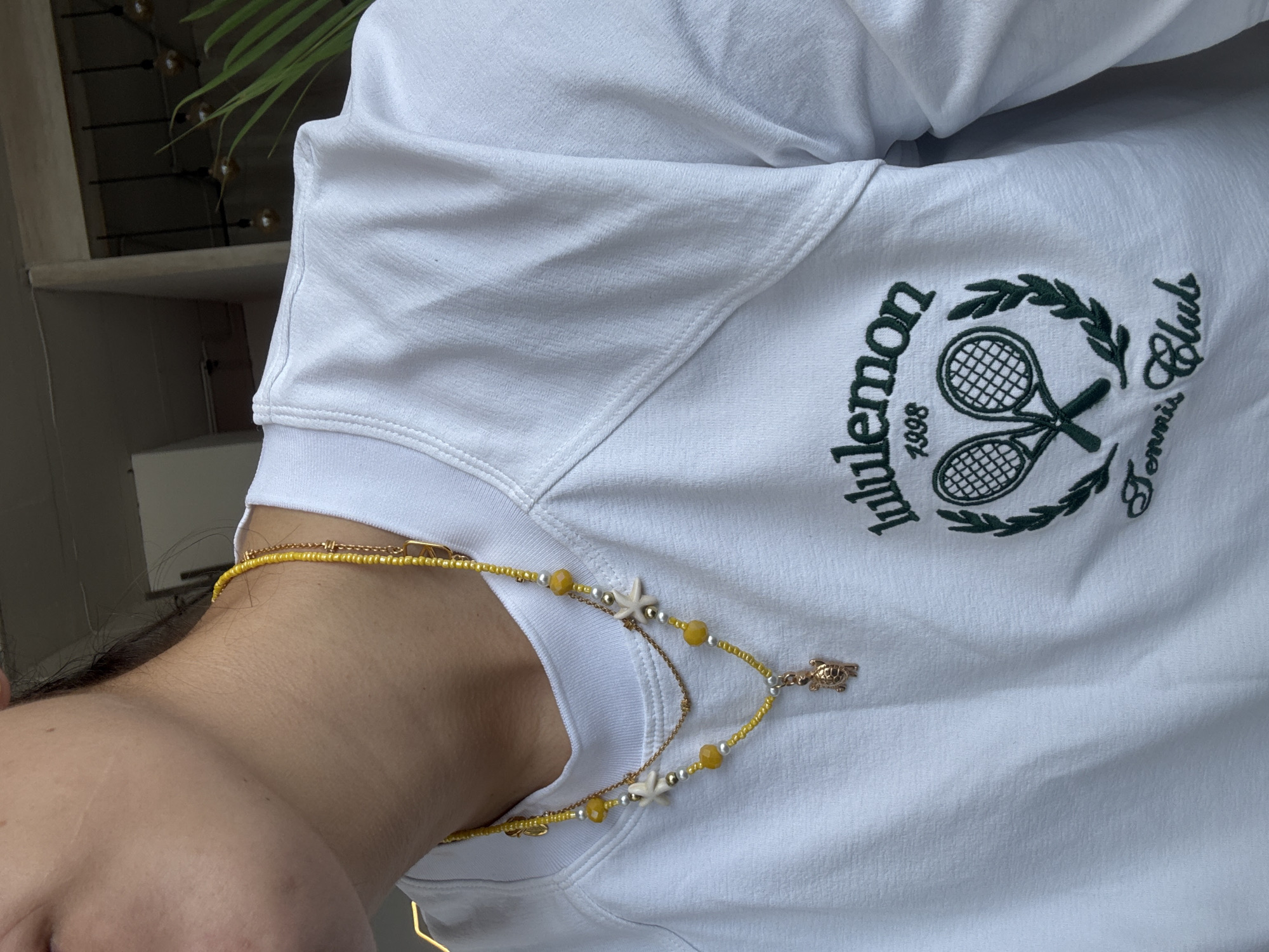 A person wearing a white Lululemon Tennis Club sweatshirt is shown in a close-up shot. A layered yellow beaded necklace with a turtle charm and starfish accents is visible around the person's neck. The photo appears to be taken indoors. It is impossible to determine if the location is in the city of Puebla, Mexico, based on the visual information provided.