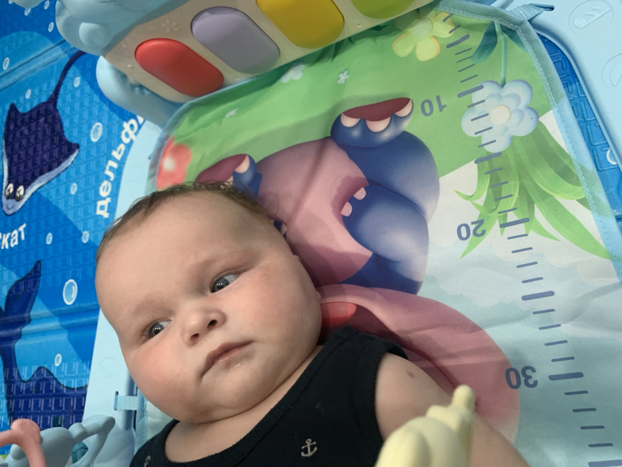 The image shows a close-up of a baby lying on its back on a playmat. The baby is looking up and to its left, with its eyes open. The playmat has various colorful illustrations, including an octopus, a stingray, and flowers. There is also a ruler printed on the mat, with markings up to 30. The baby is wearing a black onesie with a small anchor emblem. The playmat is on the floor, and a portion of a blue plastic toy is visible in the upper right corner of the image. The image appears to have been taken indoors, possibly in a nursery or playroom. No specific location in Stavropol, Russia, can be identified from the image.