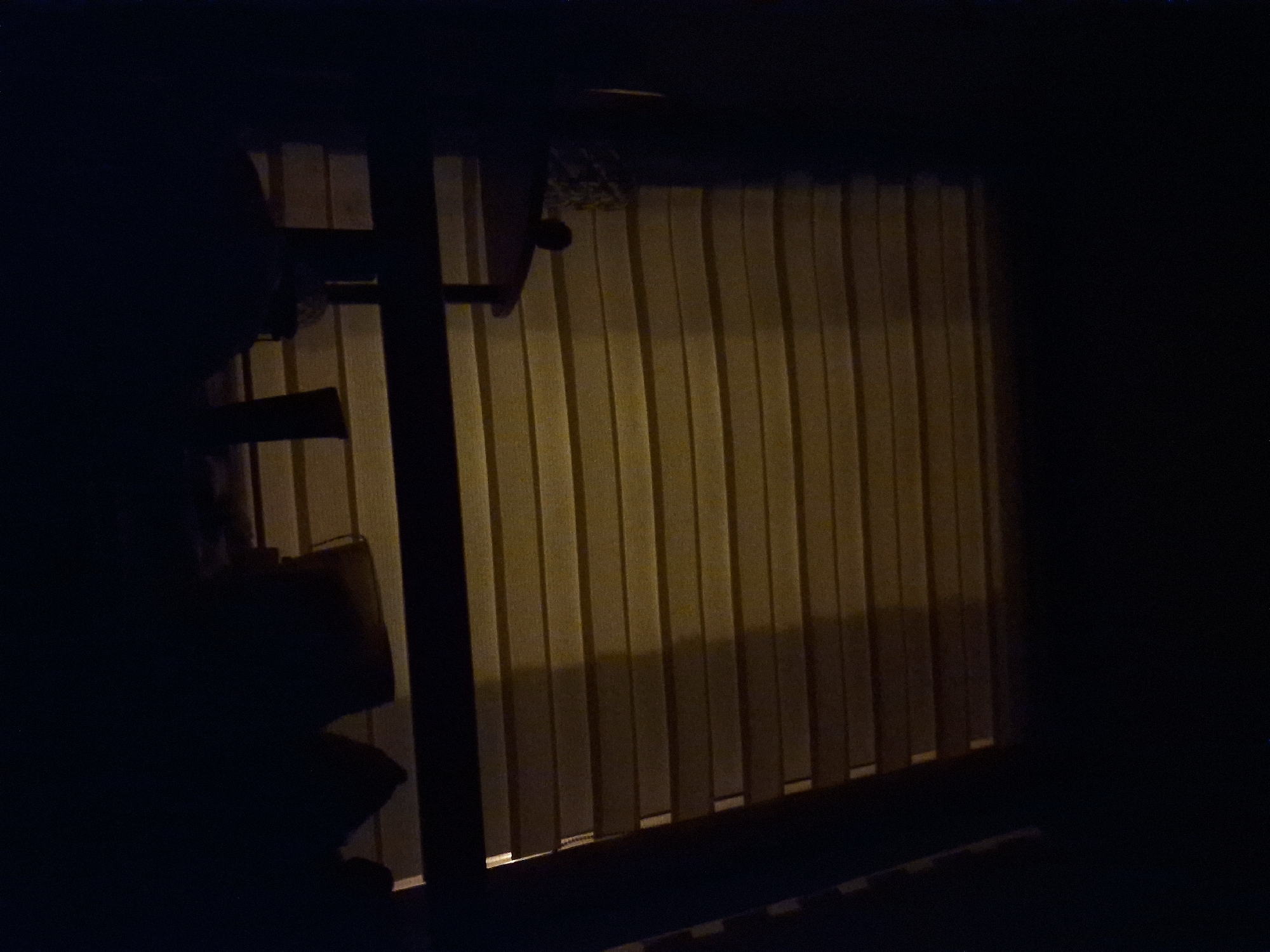 The image depicts a dimly lit indoor scene, possibly a room with blinds covering a window. A dark silhouette of a small table is visible on the left, with what appears to be a plant or decorative object on its surface. In the foreground, darker silhouettes of furniture or possibly people are present. The overall lighting suggests it is nighttime, with a warm yellow light illuminating the blinds from behind. The location is identified as Częstochowa, Poland, though no specific landmarks or urban elements are discernible to confirm this within the image itself. The scene is static and quiet.