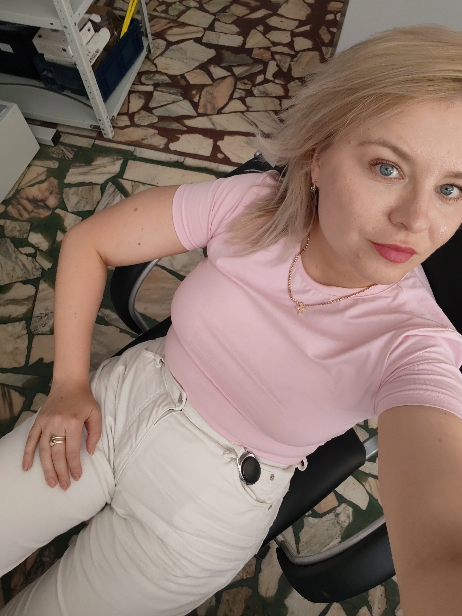 A blonde woman with blue eyes and pink lipstick is sitting in a chair, looking at the camera. She is wearing a pink t-shirt and white pants. A gold necklace with a cross pendant is visible around her neck. A ring with a stone is on her right hand. The floor is tiled with a mosaic pattern of beige, brown, and green stones. In the background, there is a white shelving unit with various items, including boxes and a blue folder. The scene appears to be indoors, possibly an office or a room with shelving. The lighting suggests it is daytime. The woman's expression is neutral. No visible text is present in the image. The location is Izhevsk, Russia.