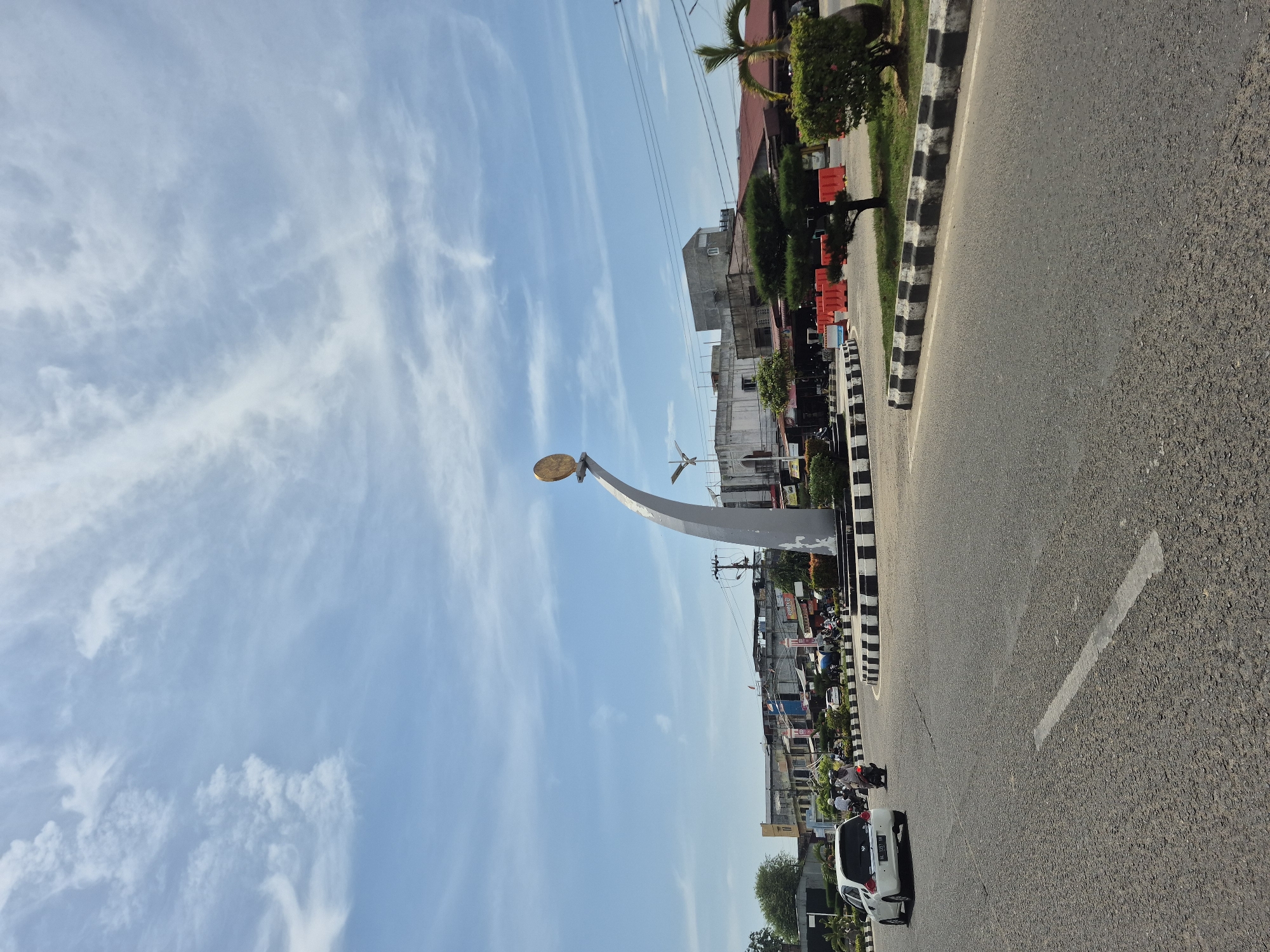 The image captures a vibrant street scene in Lhokseumawe, Indonesia, under a clear blue sky. The photo is oriented vertically, showing a wide asphalt road with a white lane marking on the right. A prominent median or sidewalk separates the road from a row of buildings, delineated by distinct black and white striped curbs.
Standing on this median is a unique white, curved monument or lamppost, topped with a golden, circular emblem, adding a distinctive local feature to the landscape. Along the median, there are patches of grass, small trees, and several orange traffic barriers.
In the mid-ground, various multi-story buildings line the street, suggesting a mix of commercial and residential establishments. Power lines are visible strung across the sky above these structures. Several vehicles are present: a white car with the license plate "BL 1621 HL" is visible towards the bottom right, and numerous motorcycles are parked or moving further down the road. A few people are indistinctly visible near the motorcycles and buildings, engaged in daily activities.
The sky is a bright, clear blue, adorned with sparse, wispy white clouds, indicating fair weather. The bright daylight suggests it is either morning or afternoon. The license plate "BL" confirms the location within Aceh province, aligning with Lhokseumawe. The overall scene depicts a typical, busy urban environment in Indonesia.