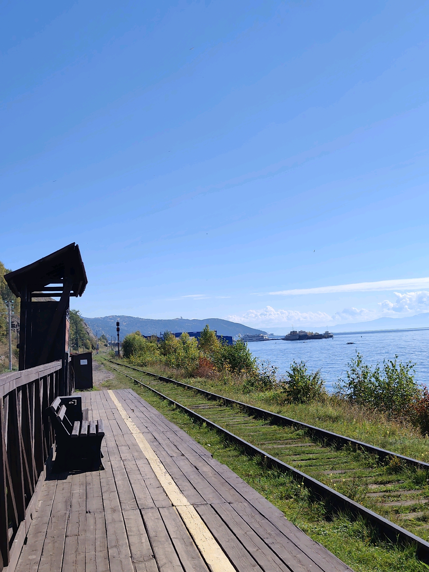 A wooden train platform with a single bench is situated beside a railway track, which runs parallel to a vast body of water. In the distance, several boats are visible on the water, and mountains form the horizon under a clear blue sky. The scene is located at a train station along the Circum-Baikal Railway in Russia.