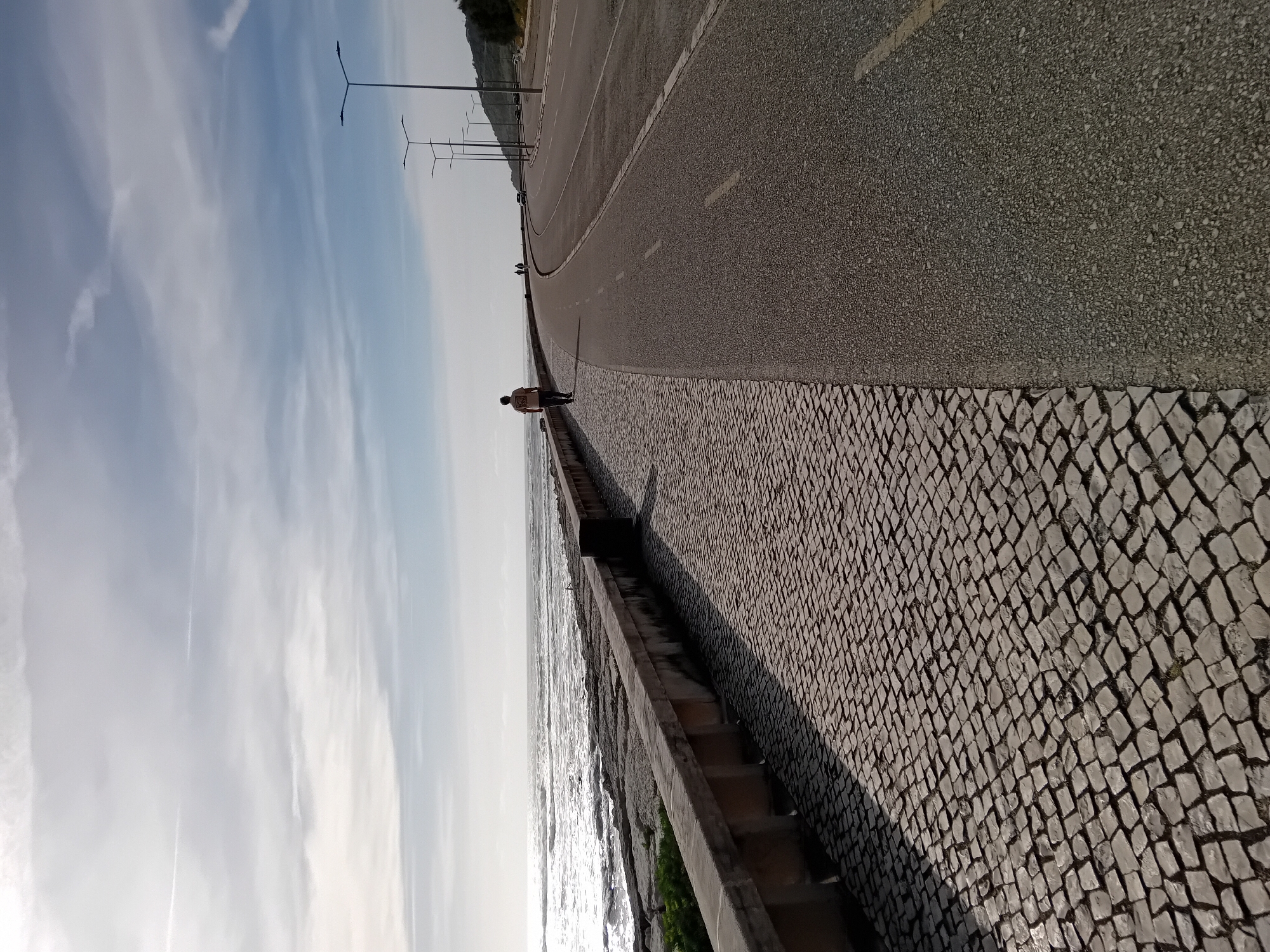 The image captures a scenic coastal promenade and road under a bright sky. The scene is oriented in a landscape format, with the ocean visible on the left side, stretching to the horizon. Waves are gently breaking near a concrete retaining wall that separates the water from a wide cobblestone promenade. The cobblestones feature a distinct diamond-shaped pattern.
A single person, seen from behind, is walking along this promenade away from the viewer, positioned closer to the concrete barrier. To the right of the promenade, a paved asphalt road curves gently into the distance. This road has a dashed white lane marker. A series of modern lampposts with horizontal arms line the far side of the road, extending into the background.
In the upper right background, a distant hillside or landmass covered with sparse vegetation is visible. The sky overhead is bright blue with scattered wispy, white clouds. The lighting suggests it is daytime, possibly late afternoon or early morning, as long shadows are cast by the lampposts and the walking person onto the promenade, indicating the sun is low in the sky, likely to the right or slightly behind the viewer. There is no visible text in the image. The overall impression is one of a calm, open outdoor space suitable for walking or driving by the sea.