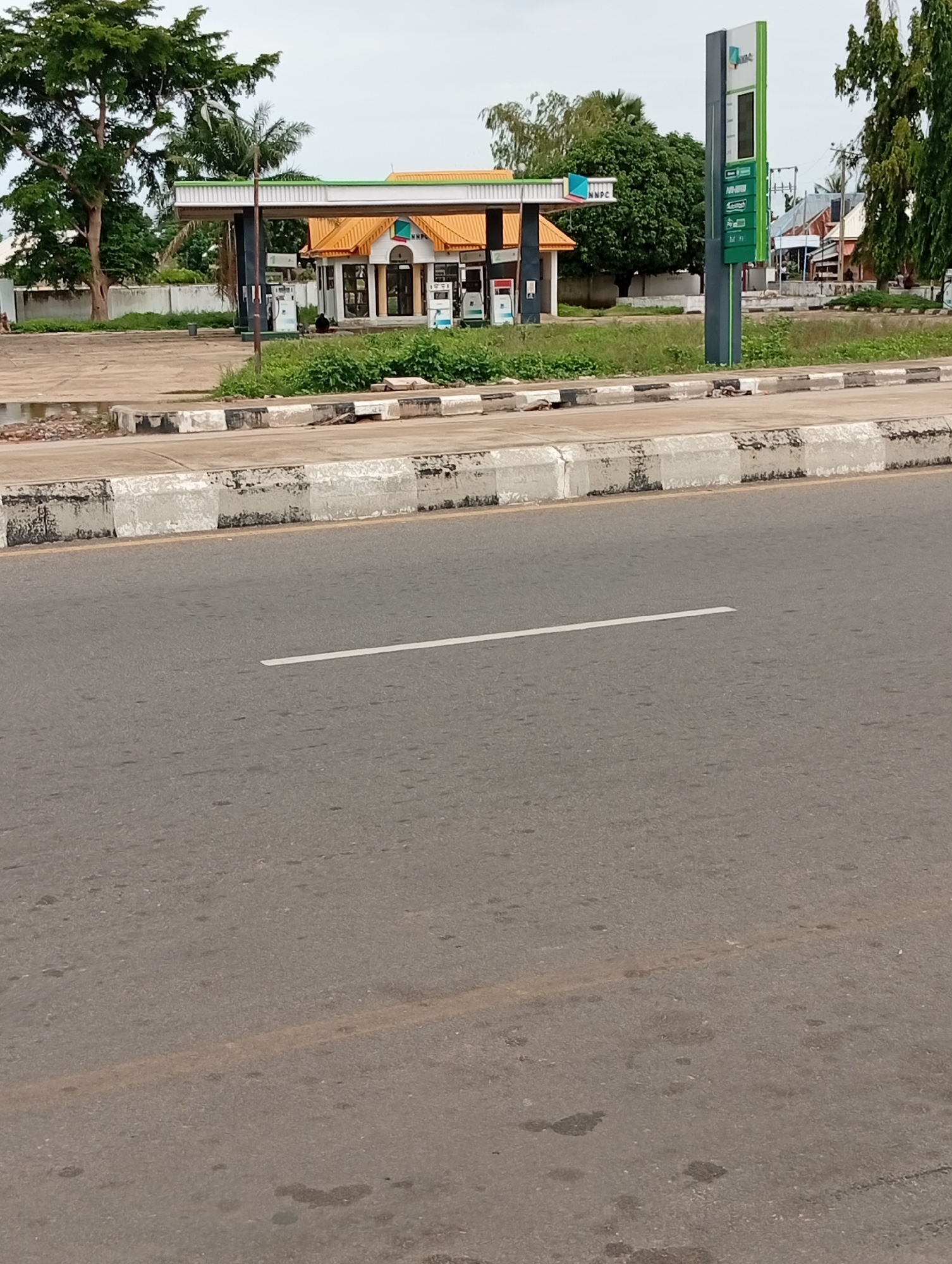 The image shows a view of a roadside NNPC gas station in Jalingo, Nigeria. In the foreground, a portion of a paved road with a white line marking is visible. Beyond the road, a concrete curb with black and white markings separates the road from the gas station area. The gas station itself features a building with an orange roof, several gas pumps, and a tall signpost with the NNPC logo and pricing information. Trees and other vegetation surround the gas station. The sky is overcast.