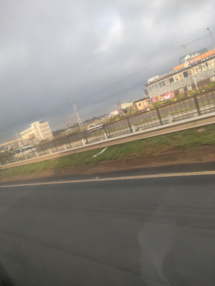 The image displays an urban roadside scene in Theta ward, Kenya, under an overcast sky. The foreground features a dark asphalt road with a faint yellow line delineating a lane. A grassy median separates this road from a concrete barrier topped with a metal guardrail. Beyond the barrier, multiple vehicles, including a white van or SUV, are visible on an adjacent thoroughfare or parking area. In the background, a two-story commercial building on the right features signage reading "RESTAURANT | CHOMA ZONE" and "TASTIES IMMING," with an additional sign for "Shaki" above. To the left, a large, light-colored multi-story building is visible. Overhead power lines traverse the frame. No individuals are discernible, and no specific actions or interactions are depicted.