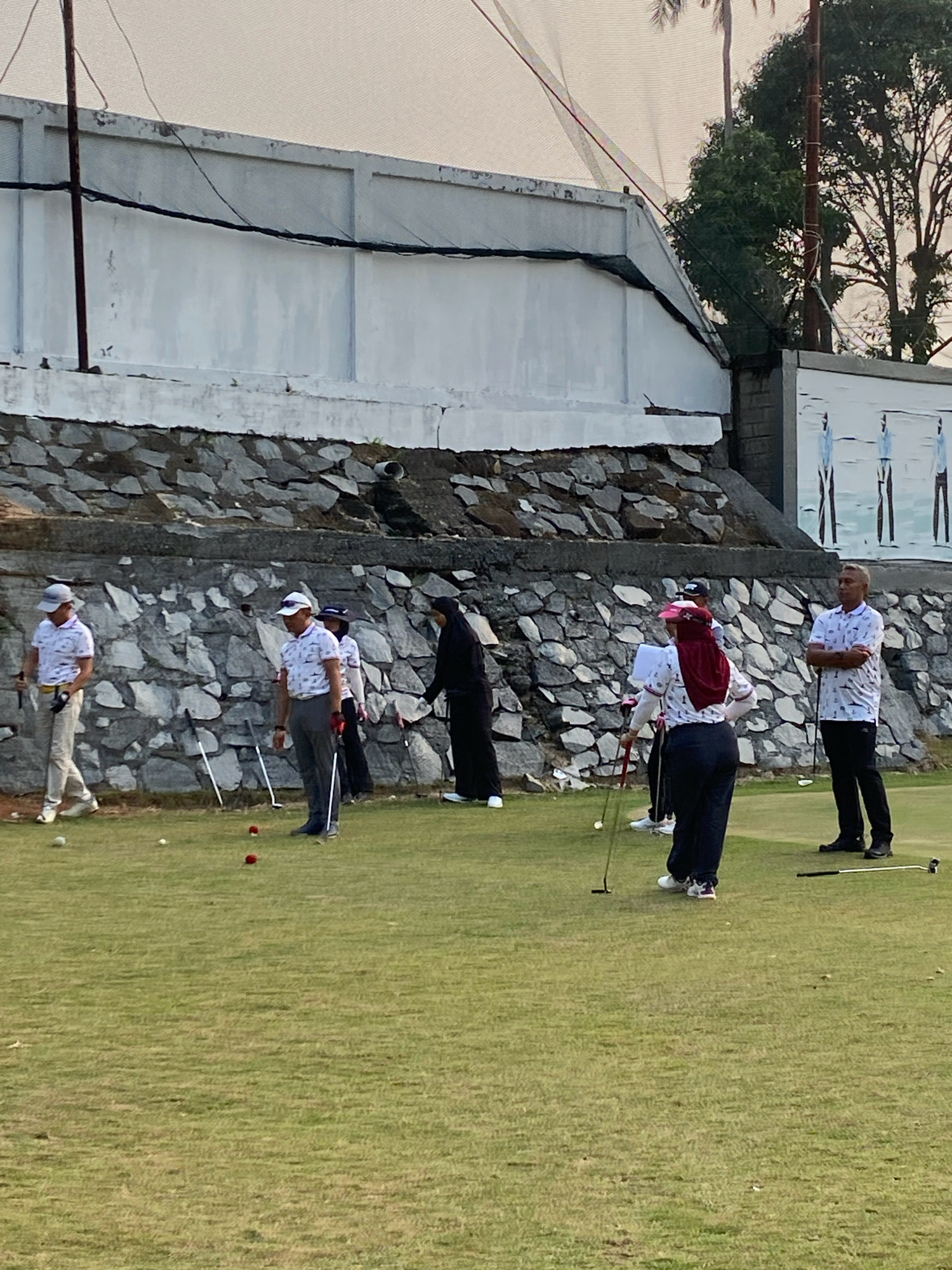 The image depicts a group of people playing golf on a grassy field, likely a golf course in Tanjung Pinang, Indonesia. The scene is outdoors, with a large concrete wall topped with rocks forming the background. A white building with a textured surface is visible above the wall, and trees can be seen further in the distance. The sky is overcast.
There are at least six individuals visible. Two men in the foreground are dressed in white polo shirts with a pattern, grey pants, and hats. One man on the left is walking, while the other on the right stands with his arms crossed. Another man, also in a white patterned polo shirt and grey pants, is standing further back and appears to be holding a golf club. A woman wearing a red hijab and a white patterned polo shirt is also present, holding a golf club. Two other individuals, one in a black abaya and hijab and another partially obscured person, are also on the field.
Several golf clubs are planted in the ground, and there are at least three golf balls visible on the grass. The people appear to be engaged in practicing or playing a game of golf. The overall impression is one of a casual recreational activity taking place on a golf course.