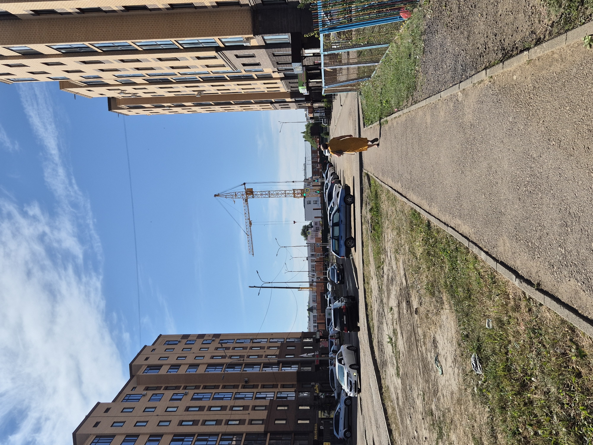 In Kokshetau, Kazakhstan, a woman in a yellow dress walks along a paved sidewalk. To her left, a modern apartment building with light brown brick facades and dark windows rises, with a parking lot filled with cars in front of it. To her right, another identical apartment building stands, adjacent to a fenced-off area with grass and dirt. In the background, a construction crane is visible against a clear blue sky with wispy clouds. The ground around the buildings is a mix of dirt, gravel, and sparse grass, with some litter present. The scene depicts a residential area with ongoing construction.