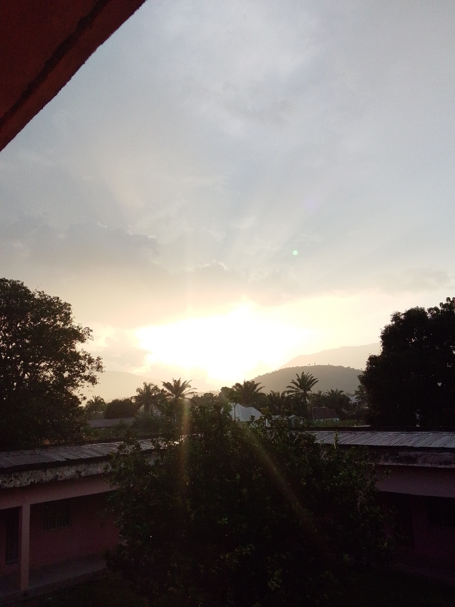 The image captures a serene sunset over a village in Serti, Nigeria. The sky is a soft blend of pale blue and warm yellow, with the sun peeking through a band of clouds, casting gentle rays across the landscape. In the foreground, the silhouettes of lush trees and the rooftops of several buildings are visible. One building, in particular, features a reddish-pink facade and a corrugated metal roof. The environment appears to be tranquil, with no people or animals visible in the frame. The overall impression is one of a peaceful evening in a rural Nigerian setting.
