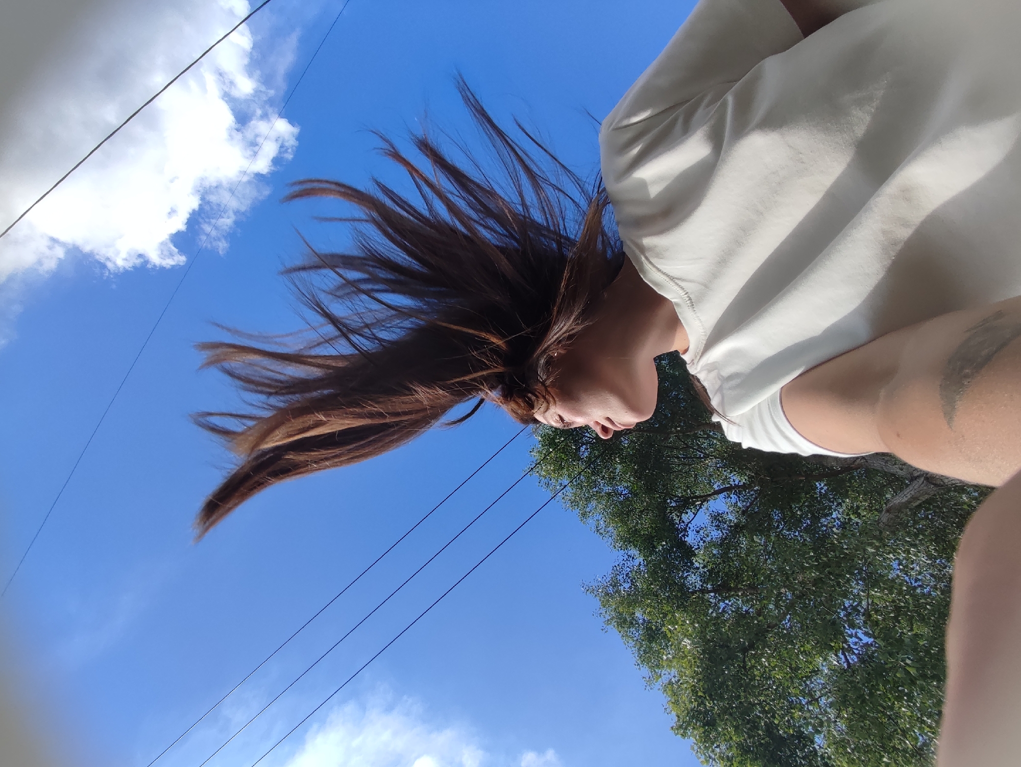 The image is taken from a low angle, looking upwards. The subject is a person with brown hair wearing a white t-shirt. The hair is spread out, suggesting movement. The background is a clear blue sky with scattered white clouds. In the distance, there is a tree with green leaves. Power lines are also visible against the sky. While I cannot ascertain the exact location within the city of Chaikovsky, Russia, based on the visual information provided, the presence of trees, power lines, and a clear sky suggests an outdoor urban environment.