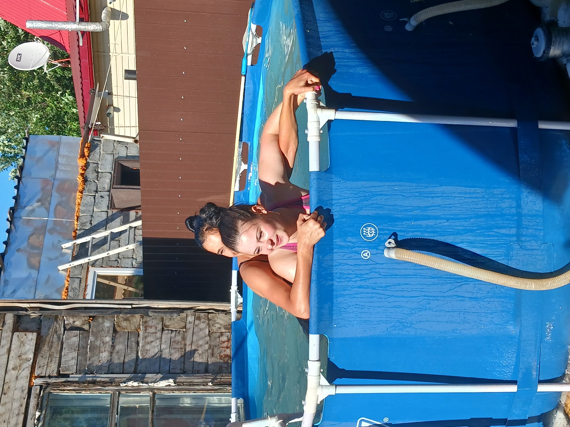 In the city of Chik, Russia, two young women are enjoying a sunny day in a blue inflatable swimming pool. The pool is set up outdoors, with a rustic wooden building and a brown metal fence visible in the background. One woman, in a pink bikini, is leaning on the edge of the pool, her arms resting on it, while the other woman, in a dark swimsuit, embraces her from behind. The water is clear, and the sun casts shadows on the pool's exterior. The scene appears to be a casual, outdoor leisure activity in a residential area.