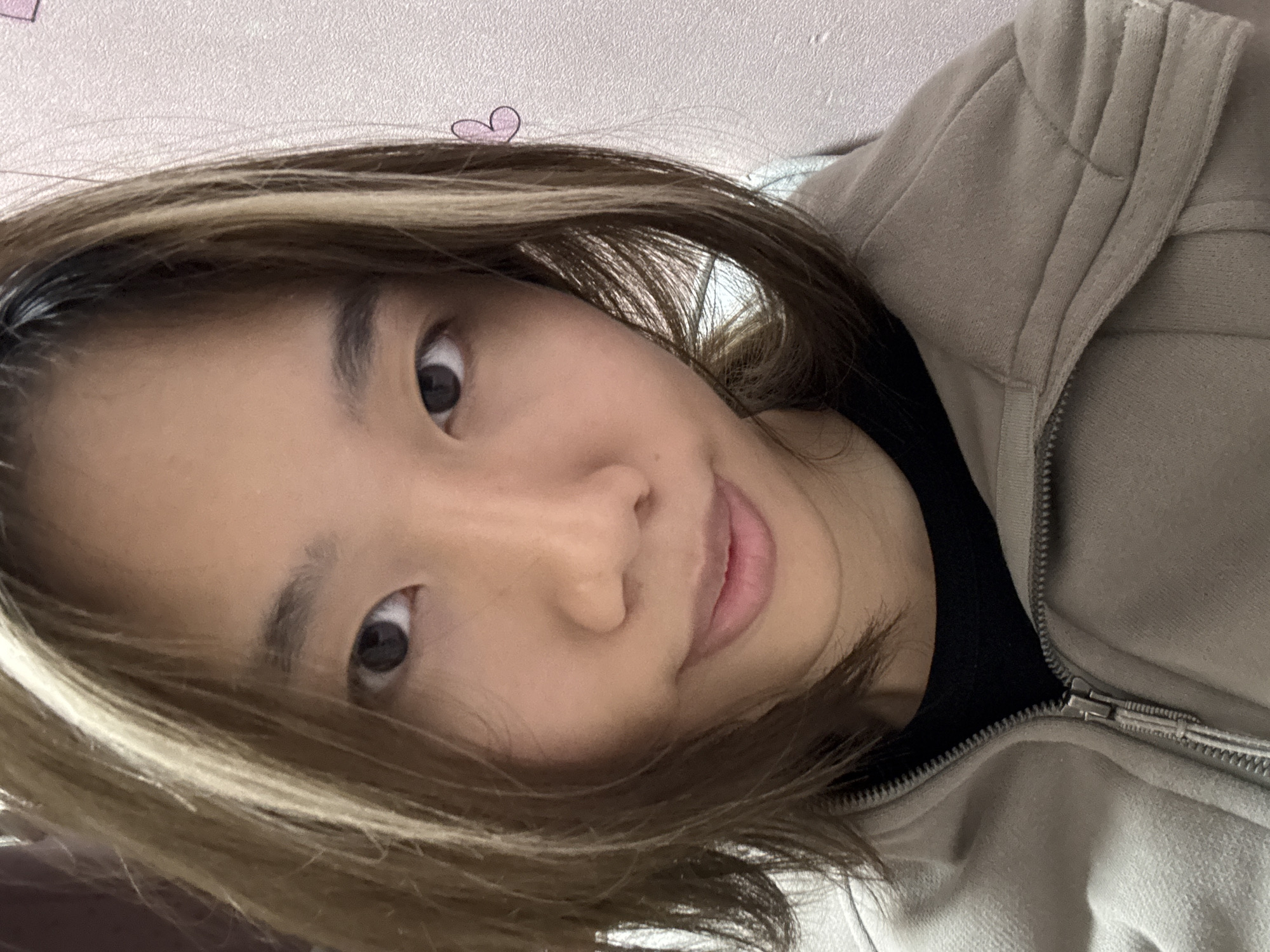 A person with brown hair and blonde highlights is looking at the camera with a slight smile. They are wearing a beige jacket with a black shirt underneath. A small pink heart is drawn on the wall behind them. The location, Shapingba District, China, cannot be verified from the image.