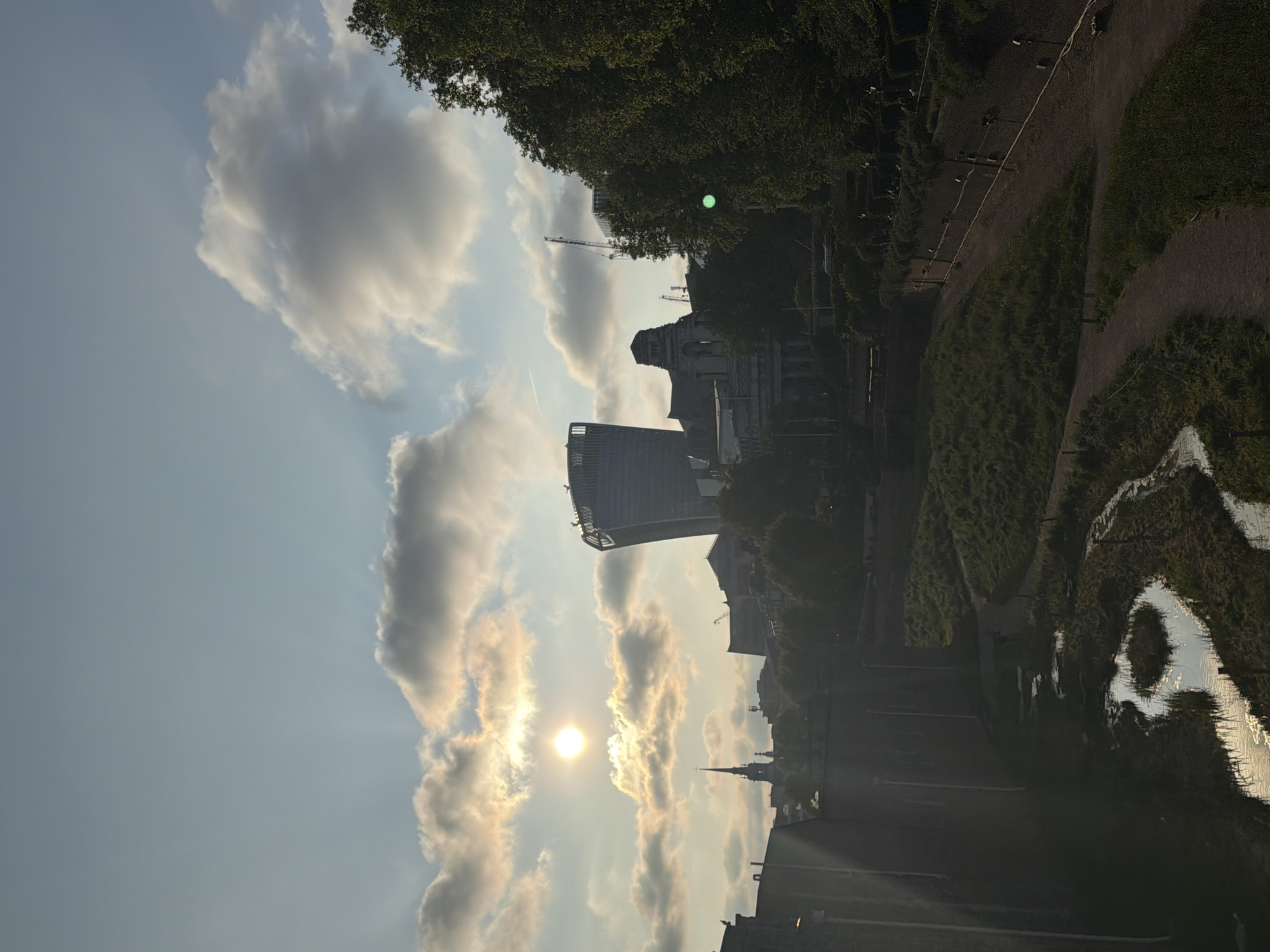 The image depicts a cityscape in London, United Kingdom, under a sky filled with clouds and the setting sun. Dominating the scene is the "Walkie Talkie" building (20 Fenchurch Street) and a view towards the city with the distinctive spire of Christ Church, Spitalfields. To the side, there appears to be a park or green space, possibly with a water feature, visible to the side of the frame. The image captures a sunset or late afternoon light, giving the clouds a golden hue. The location seems to be a vantage point overlooking a portion of the city skyline.