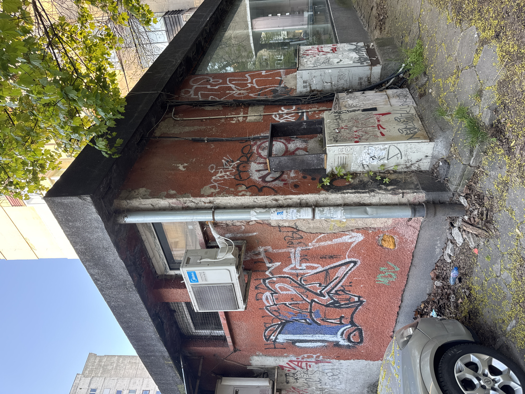 The image presents an upright view of a neglected urban building exterior and its surroundings in Sofia, Bulgaria, captured on a sunny day. The focal point is a reddish-brown textured wall, heavily covered in various graffiti tags and abstract designs in white, black, blue, and red spray paint. Above, a large, dark-framed reflective window is visible, with a prominent dark concrete or stone overhang extending across the building's facade.
Several utility installations are mounted on the wall. Multiple black electrical conduits and silver pipes run along its surface. Two air conditioning units are visible; the upper one clearly displays the "Midea" brand logo in blue, along with a partially legible phone number "088 8 XX XX 88" and some Cyrillic text. Below the window, several metal utility boxes are affixed, also defaced with graffiti and stickers. A small, open black box is positioned centrally on the wall.
The ground adjacent to the building is a mix of irregular paving stones and exposed earth, covered with fallen yellow pollen or petals. Patches of green grass and weeds grow between the stones and at the base of the wall. Debris such as dried leaves, dirt, and a discarded blue plastic bottle are scattered on the ground. To the bottom right, the front right quarter of a silver car, including its wheel, tire, and headlight, is partially visible, also dusted with yellow pollen. In the upper left, vibrant green leaves of a tree partially obscure the view, indicating natural vegetation in the urban setting. The bright lighting suggests it is daytime with a clear, blue sky visible in the background.