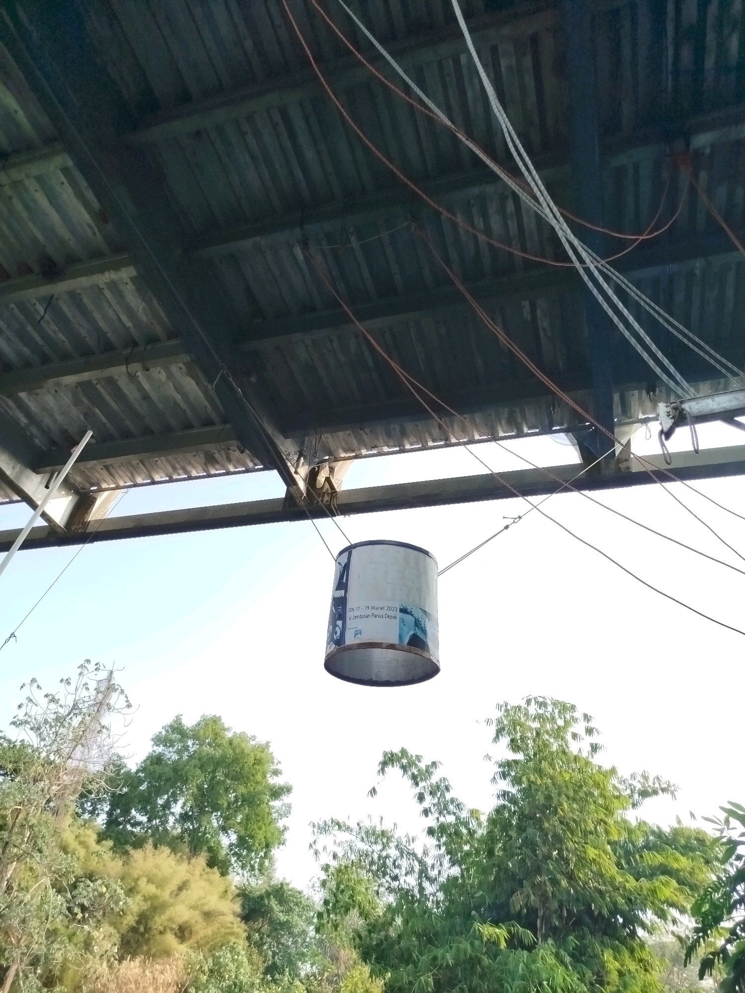 A cylindrical object, possibly a paint can, hangs suspended by wires from an overhead structure composed of corrugated metal roofing and metal beams. The object has text printed on its side, including "17 - 19 Maret 2023" and "Jembatan Panus Depok," suggesting it is related to an event or location in Depok, Indonesia. The background consists of a bright, hazy sky and lush green trees. The scene appears to be outdoors, possibly in a shaded area under a covered structure.