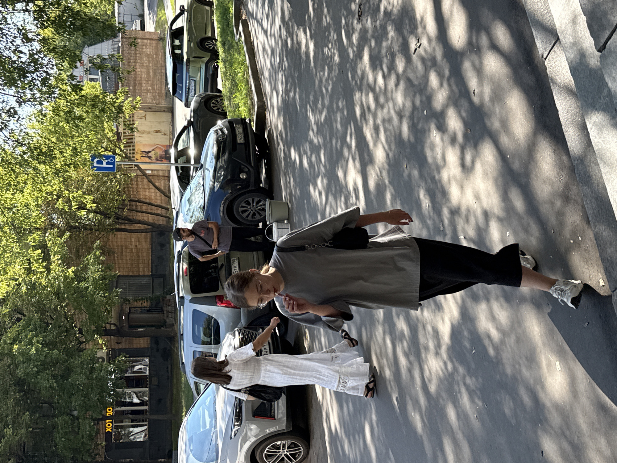 On a sunny day, with dappled light filtering through trees onto a paved area in Moscow, Russia, a young woman with glasses and brown hair, wearing a grey t-shirt and dark pants, walks forward, holding what appears to be a cigarette to her lips. Behind her, a woman in a long white dress walks away from the camera. Several cars are parked on the street, including a silver SUV, a black SUV, and a light green SUV. A man in a grey t-shirt stands near the black SUV, looking towards the right. A blue parking sign with an accessibility symbol is visible in the background. The environment suggests a residential or commercial street with trees and buildings.