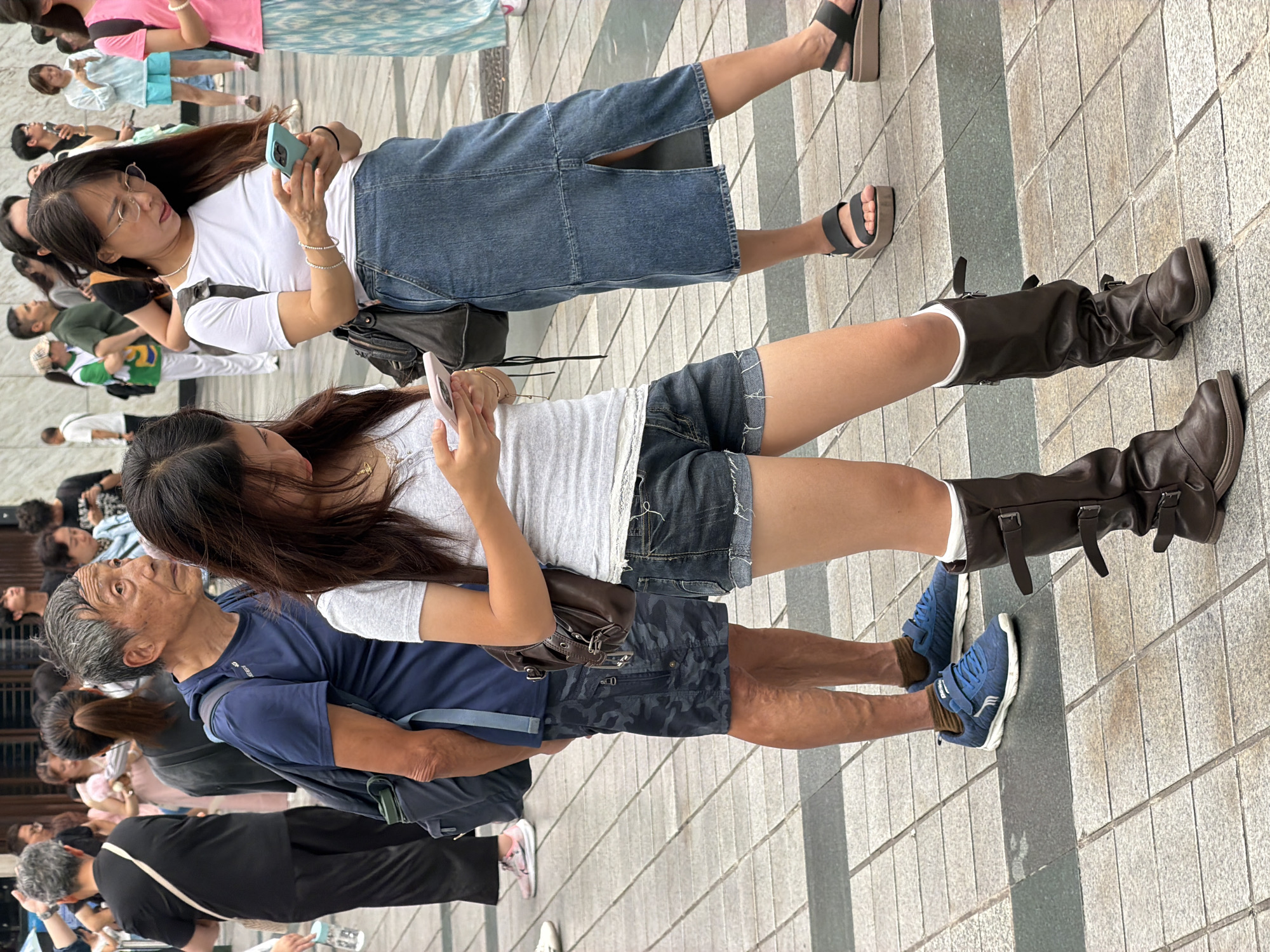 In Yuzhong District, China, a diverse group of people are observed on a tiled pedestrian walkway. In the foreground, a young woman with long dark hair, wearing a grey t-shirt and denim shorts, is engrossed in her smartphone. To her left, another young woman, also with dark hair, is looking at her phone, dressed in a white t-shirt and a denim skirt with a high slit. An older man with grey hair, wearing a blue t-shirt and camouflage shorts, stands between them, looking to his right, with a backpack slung over his shoulder. In the background, several other individuals are visible, some also looking at their phones, while others are walking or standing, suggesting a public gathering or a busy street. The environment appears to be an outdoor urban setting with a paved ground surface.
