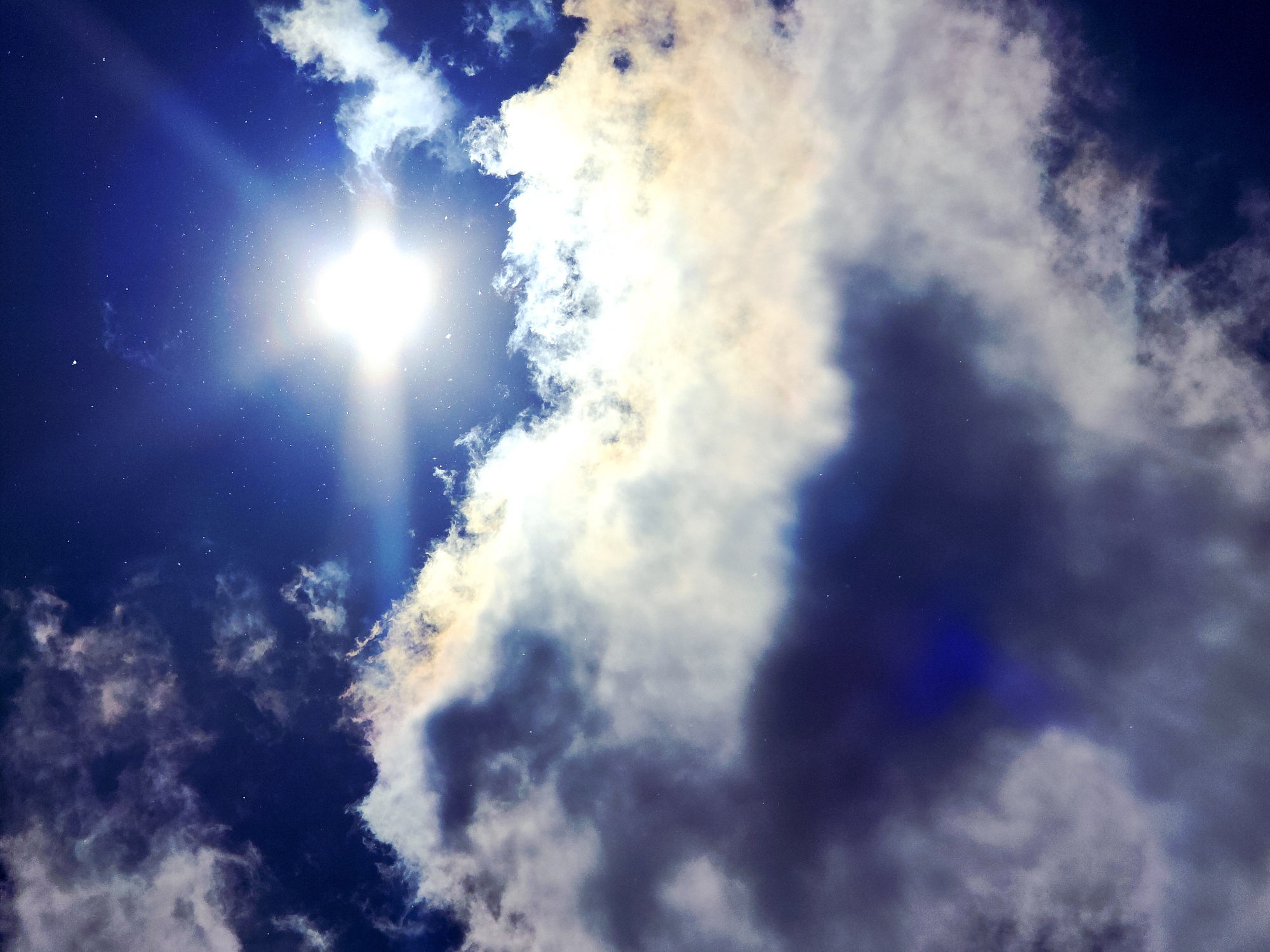 This image presents a dynamic sky scene, looking upwards towards a brilliant sun amidst varied cloud formations. The sun, located in the upper-left quadrant, emits an intense white light with prominent starburst rays and blue lens flares.
The sky is a deep, rich blue, providing a stark contrast to the illuminated clouds. A large, dense cloud formation occupies much of the right side and extends towards the center and bottom. These clouds display a spectrum of tones, from luminous white and cream where directly hit by sunlight, to subtle yellows, oranges, and hints of brown, likely from sunlight interacting with the cloud particles. Darker grey and deep blue shadows are visible within the thicker cloud masses, suggesting depth.
Smaller, wispy clouds are scattered across the blue expanse. Numerous tiny white specks are visible throughout the image, especially against the darker blue sky and within the sun's glare, which could be dust on the camera lens or atmospheric particles. The scene depicts a bright, partially cloudy day. There is no specific activity or event taking place, nor are there any discernible location cues or visible text.