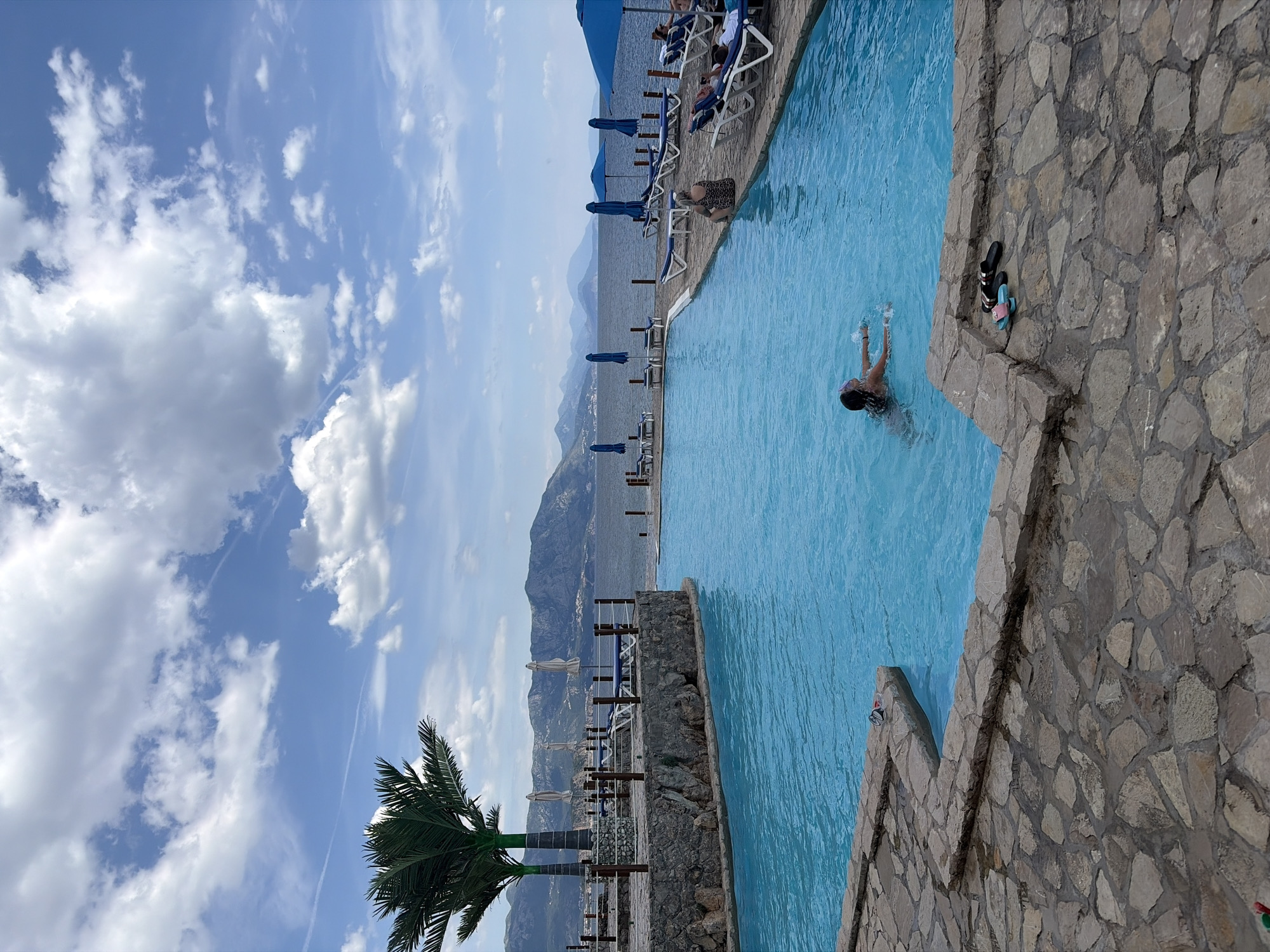 A person is swimming in a light blue swimming pool that is situated next to a stone barrier. Shoes are placed on the barrier. In the background, there are sun loungers and umbrellas, which are located close to mountains and water. A palm tree is in the foreground. The sky is blue with puffy white clouds. The location of the image is in Prijevor, Montenegro.