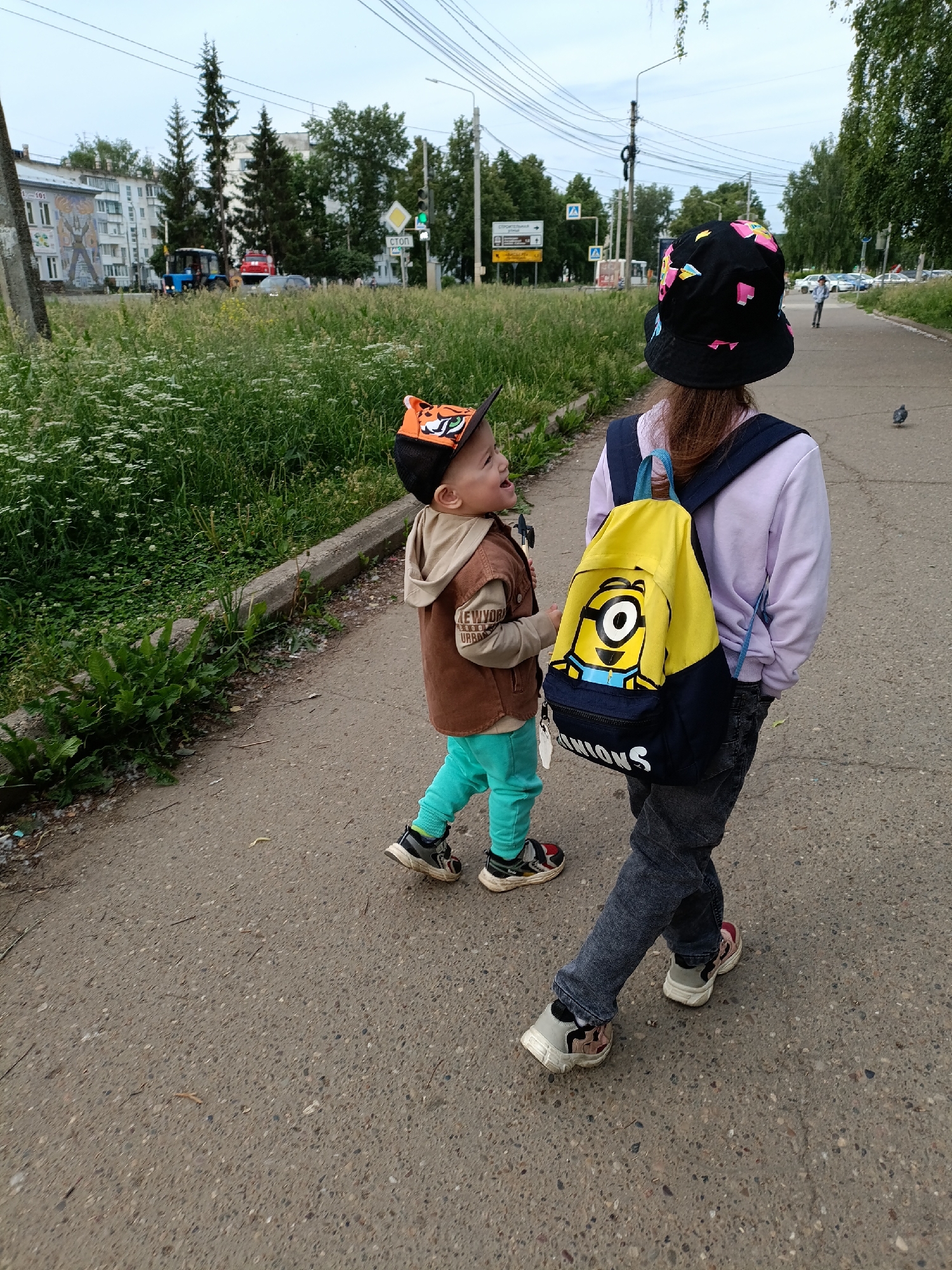 Two children are walking along a paved sidewalk next to a grassy area in Chaikovsky, Russia. The older child, seen from behind, wears a black bucket hat with colorful geometric shapes, a light purple hoodie, dark jeans, and sneakers. They carry a yellow and blue backpack featuring a large Minion character, with the word "MINIONS" visible at the bottom. The younger child walks slightly behind and to the left, looking up and smiling. This child wears an orange and black tiger-themed baseball cap, a brown and beige hooded jacket with "NEW YORK URBAN" partially visible on a patch, light blue pants, and sneakers.
The scene appears to be daytime, likely spring or summer, with an overcast sky. The sidewalk is lined with green grass and wildflowers. In the background, beyond the grassy strip, there's a road with vehicles including a blue tractor and a red truck (possibly a fire engine). Multi-story apartment buildings are visible further back, one featuring a large, colorful mosaic mural. Utility poles and power lines crisscross the sky. Several street signs are visible, including a pedestrian crossing sign and a white sign with the Cyrillic word "СТОП" (STOP) next to a green traffic light. A pigeon is perched on the sidewalk to the right.