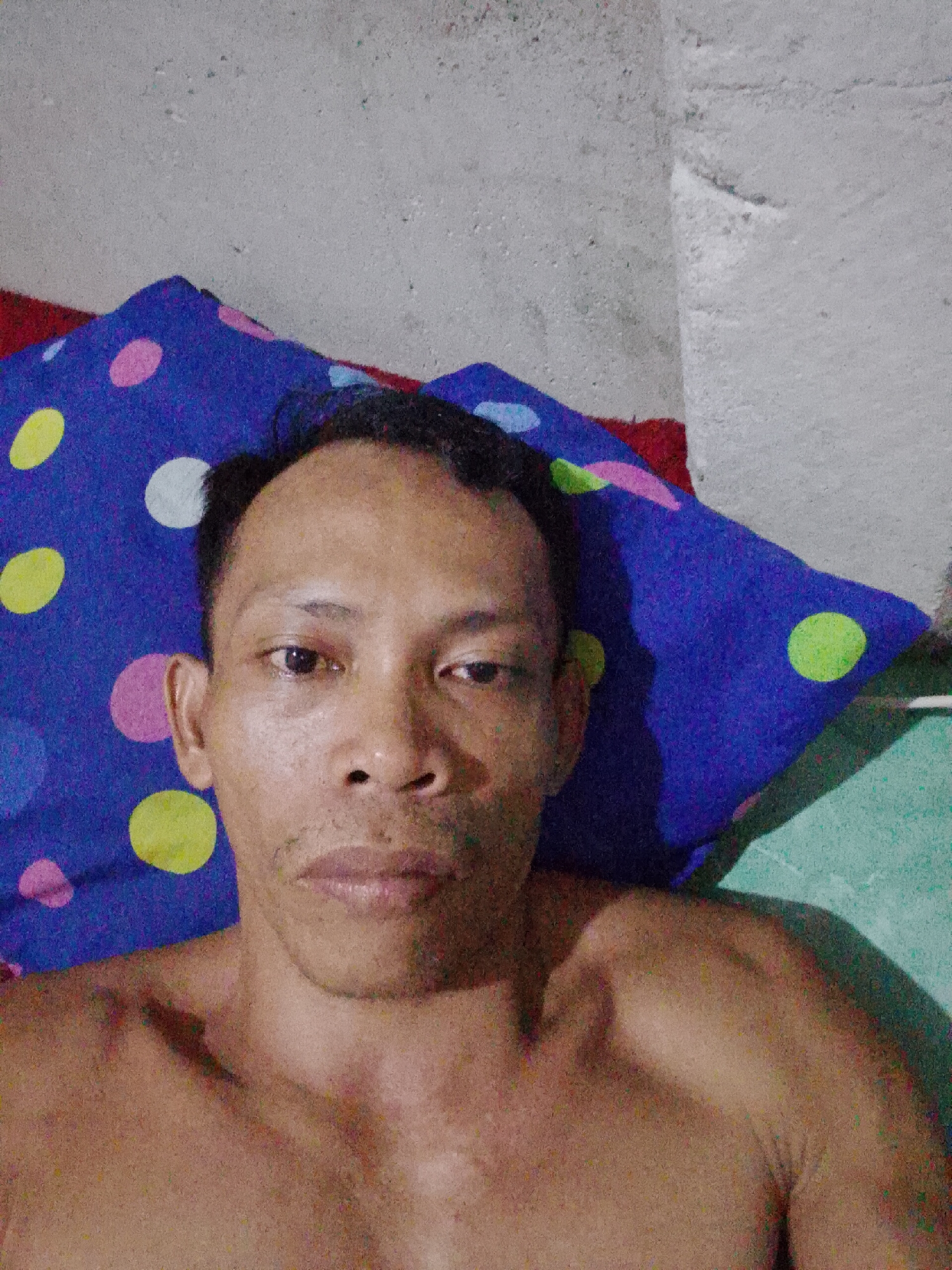 An adult man is lying on a blue pillow with pink, white, yellow, and green polka dots. The man has short, dark hair, dark eyes, and appears to be shirtless. He is in a room with a pale grey wall. The environment suggests a bedroom or living space. The location is Bukit Ketok, Indonesia.