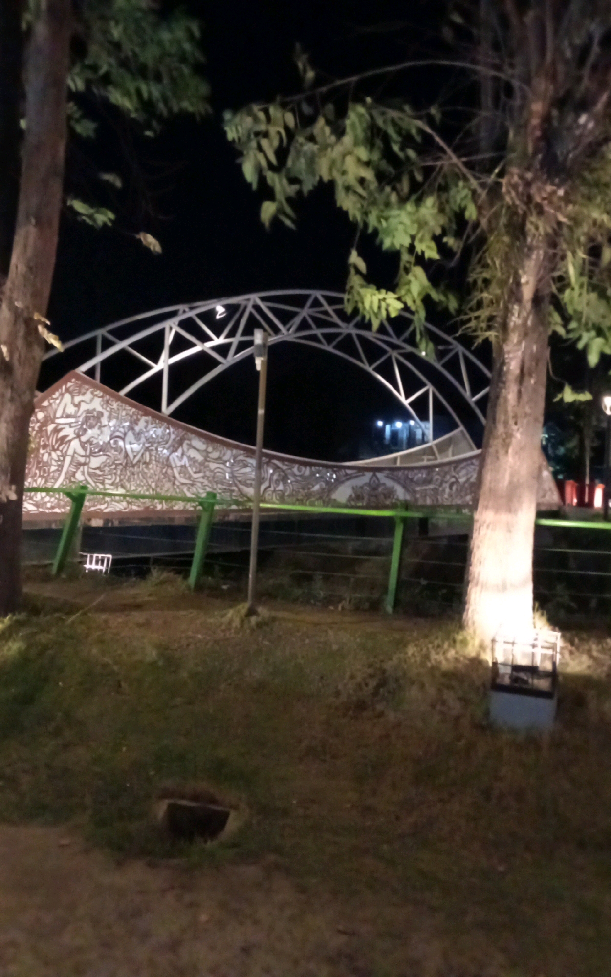 The image depicts a nighttime scene in the city of Ngasem, Indonesia, featuring an illuminated artistic structure with intricate carvings. Two trees flank the structure, their trunks illuminated by spotlights. The structure itself is a curved wall adorned with detailed carvings, set against a backdrop of a metal archway. Green railings run along the front of the structure, with a lamppost visible on the right. The ground in the foreground is covered with grass and dirt, with a dark square object on the right side of the frame. The overall atmosphere is serene and artistic, illuminated by artificial lighting.