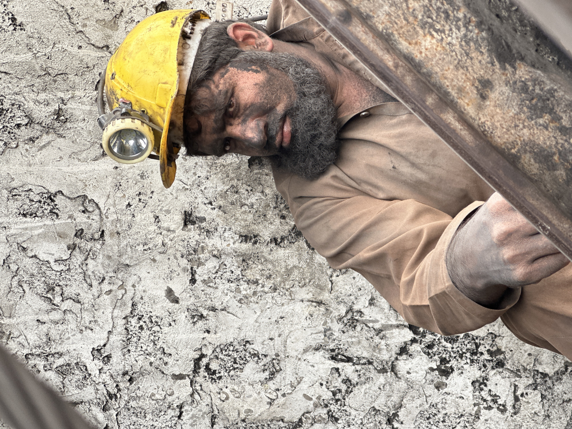 A close-up, eye-level shot shows a coal miner in Quetta, Pakistan, covered in soot. He is wearing a yellow hard hat with a headlamp, a tan long-sleeved shirt, and has a full beard. The miner's face is smudged with black coal dust, obscuring much of his features, but his eyes are visible, looking off to the left. His right hand rests on what appears to be a piece of rusted metal. The background is a textured, pale wall with dark, irregular markings, possibly the interior of a mine or a rough-hewn structure. The lighting highlights the miner's face and the texture of the background, suggesting an indoor or underground setting.