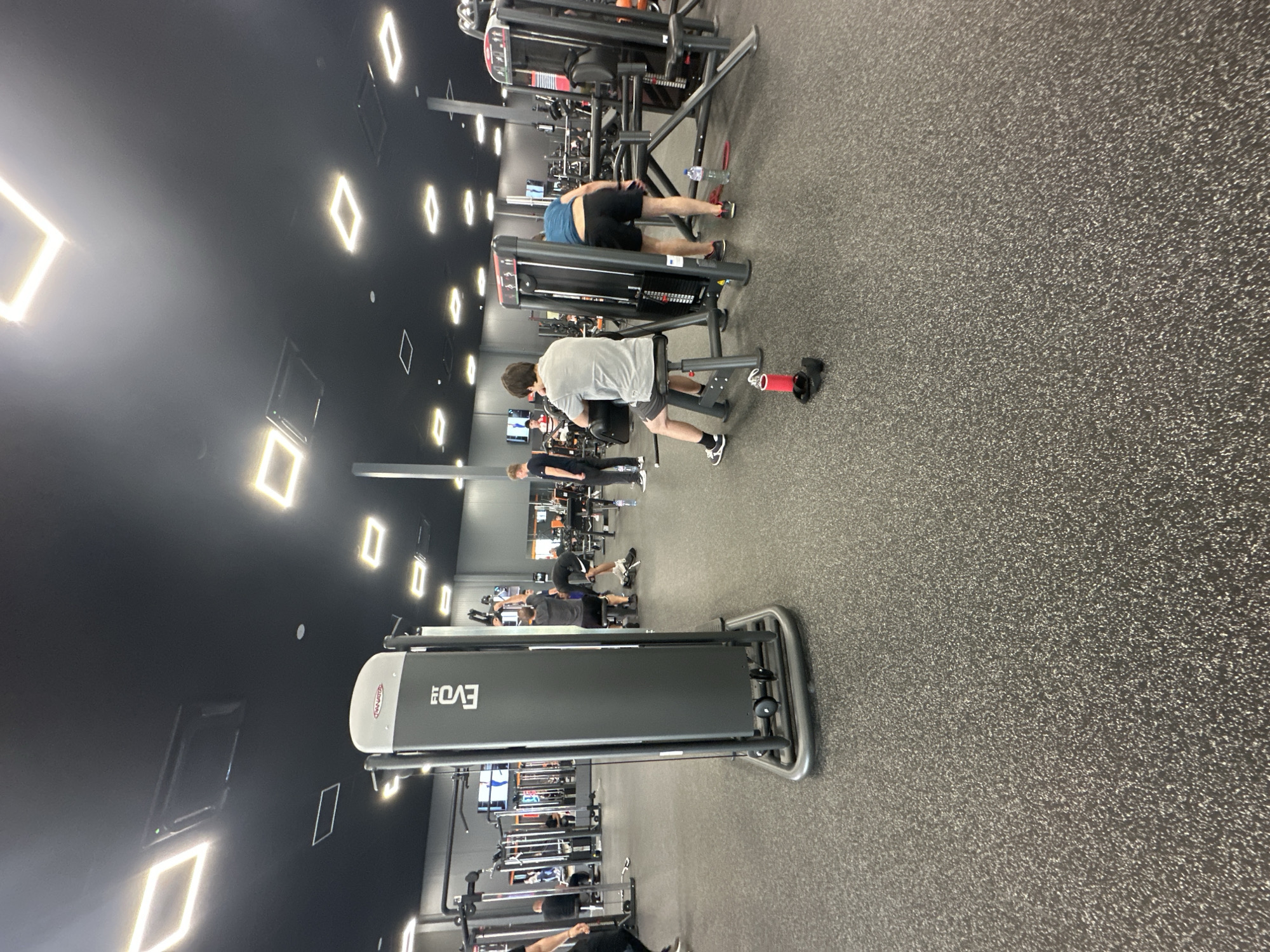 The image shows the interior of a gym. Several people are using various exercise machines. There are treadmills and weight machines. The lighting consists of rectangular, diamond-shaped lights on a black ceiling. The floor is gray. The location is unidentified regarding the city of Plovdiv, Bulgaria.