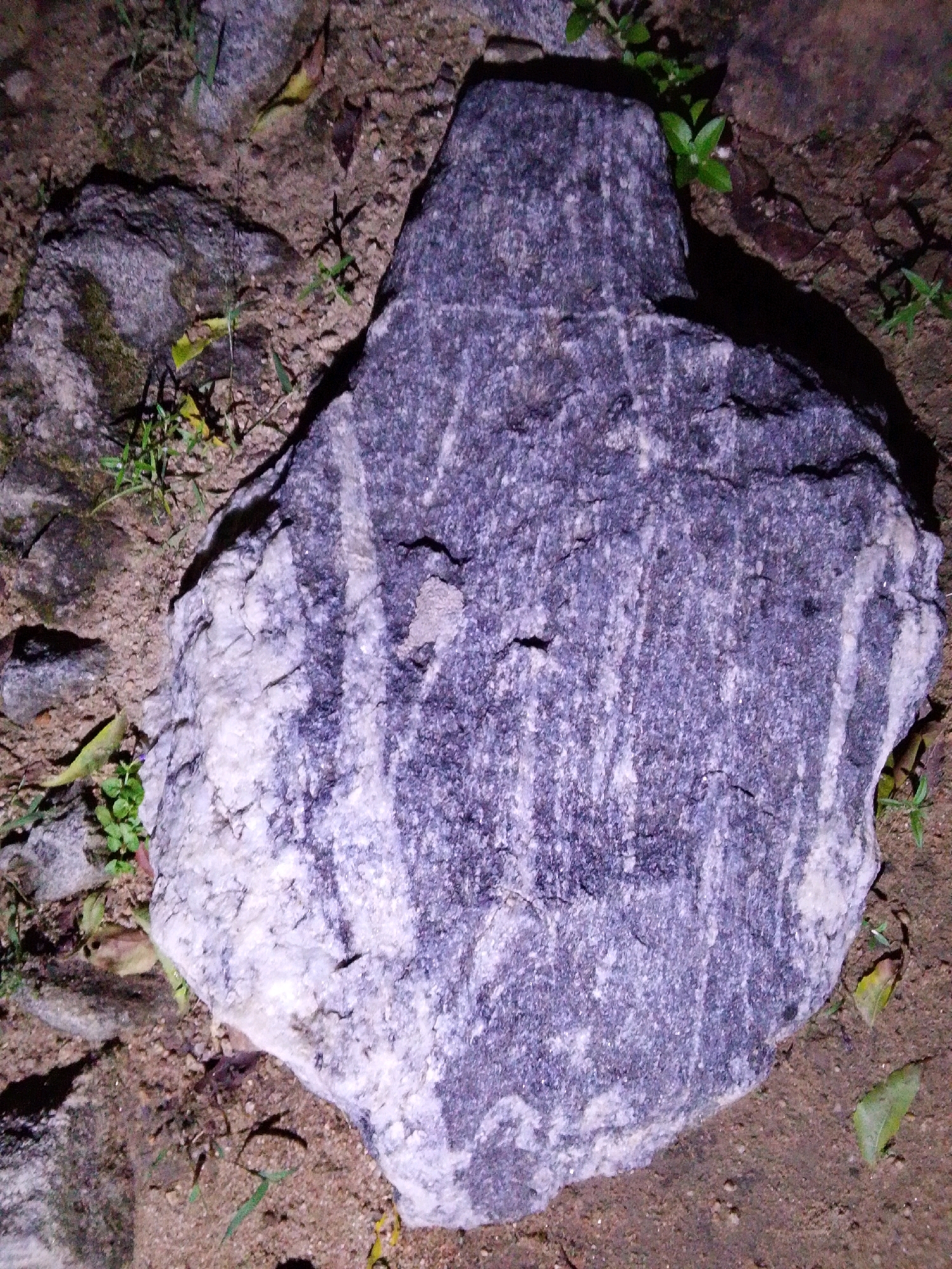 A large, dark gray and white mottled rock with distinct vertical striations is the central focus of the image. It rests on sandy, brown soil amidst sparse green vegetation and a few fallen leaves. The rock is irregular in shape with a pointed tip on the left and a rounded, uneven base. The surrounding environment appears natural, possibly a park or unpaved area, with other smaller rocks visible in the background. The lighting suggests it may be evening or a dimly lit area. No people or specific Nigerian landmarks are visible.