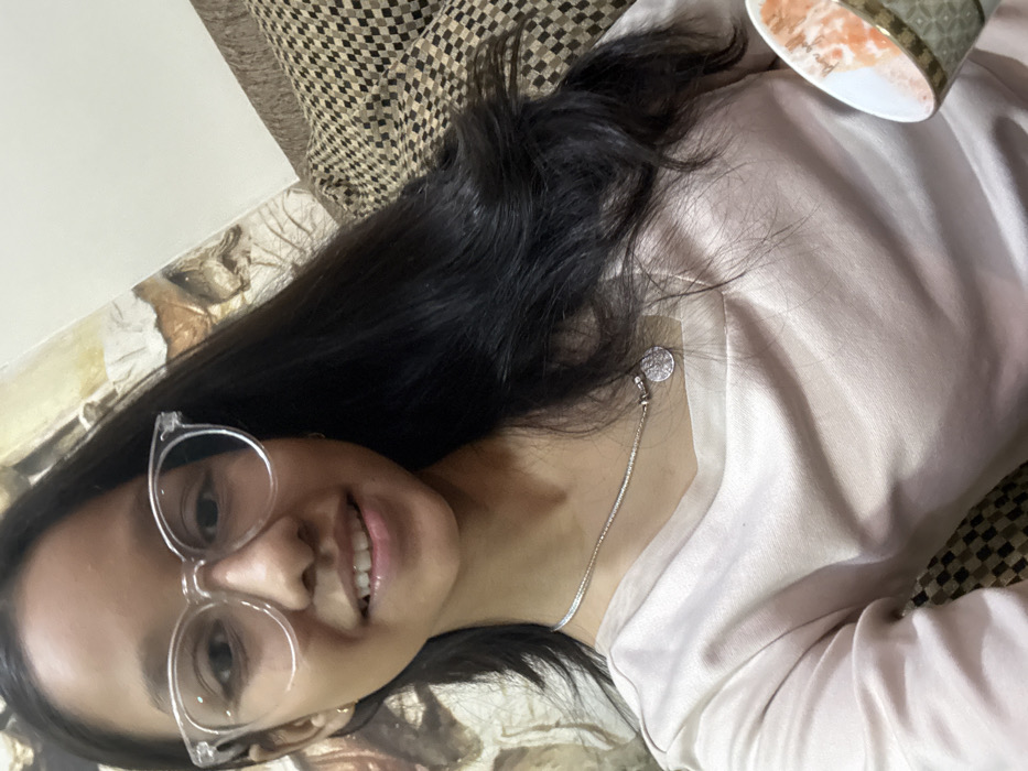A woman with glasses lies on a patterned surface. She has long, dark hair, a silver necklace, and a light pink satin top. Part of a cup is visible in the top right corner. The woman smiles towards the camera.