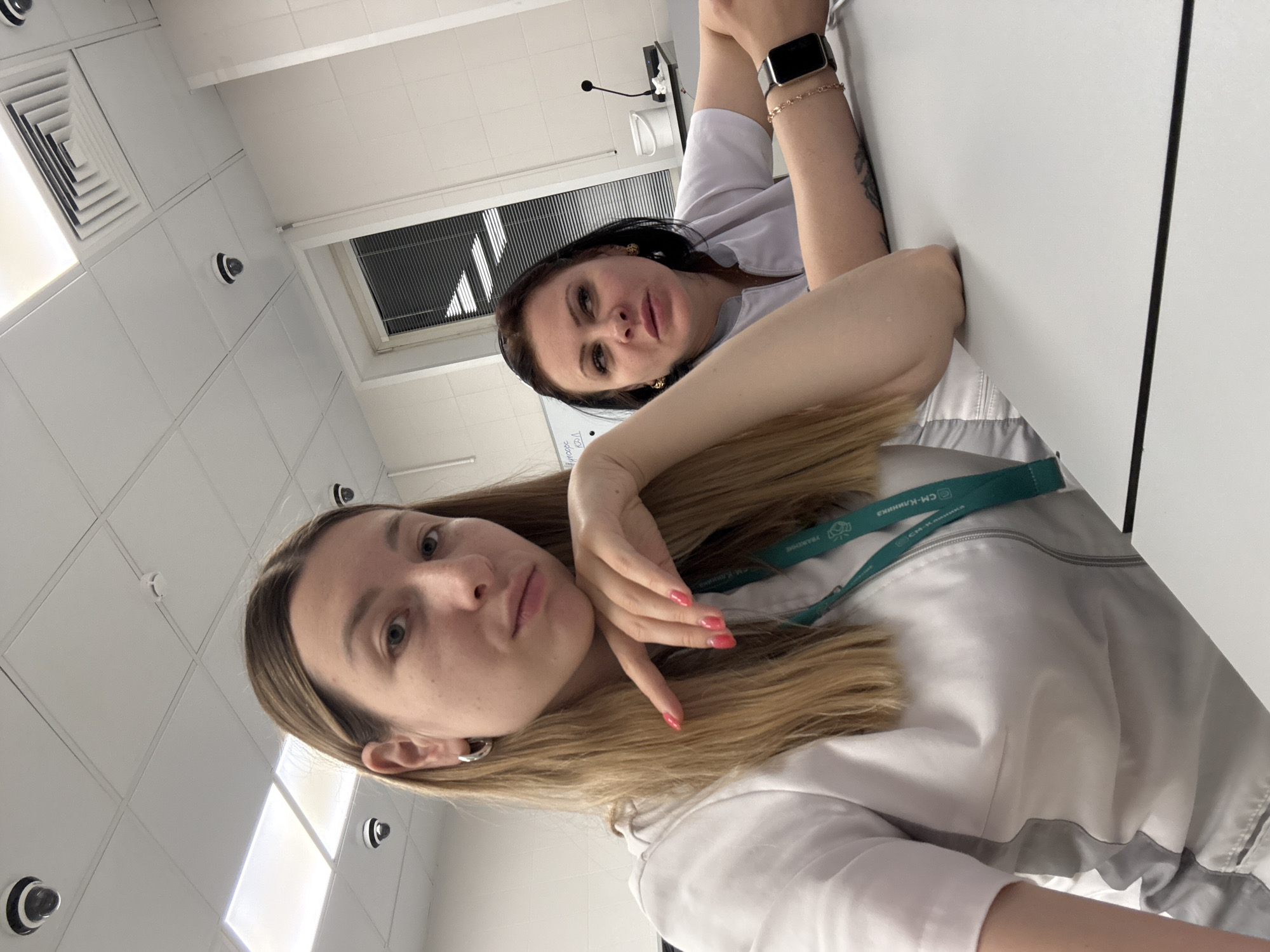 This image shows two women sitting at a desk in a brightly lit room with a white tiled ceiling. The woman in the foreground, on the left, is facing the camera and has long blonde hair, silver hoop earrings, and is wearing a white lab coat with a teal lanyard. Her right hand is resting on her chin. The woman in the background, on the right, has dark hair and is wearing a white lab coat. She is looking to her left, and her right arm is resting on the desk, displaying a smartwatch and a bracelet. Several security cameras are visible on the ceiling, and there is a window with blinds behind the woman in the background. The room appears to be an indoor, possibly institutional, setting like a classroom or laboratory. There is no visible indication of the city being Moscow, Russia, or any specific location.